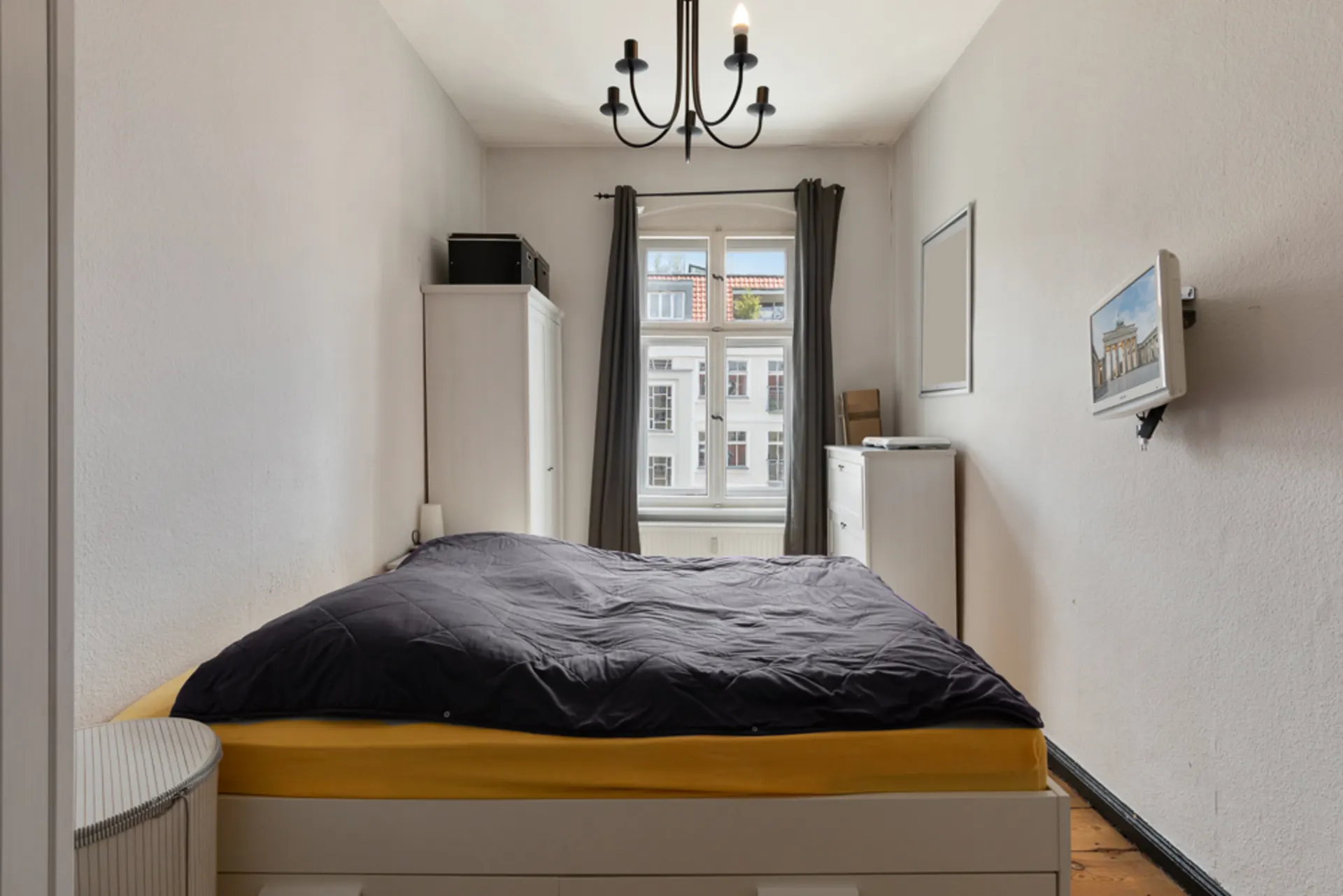 Schlafzimmer mit Doppelbett, Kleiderschrank, Fenster mit Vorhängen und an der Wand montiertem Fernseher.