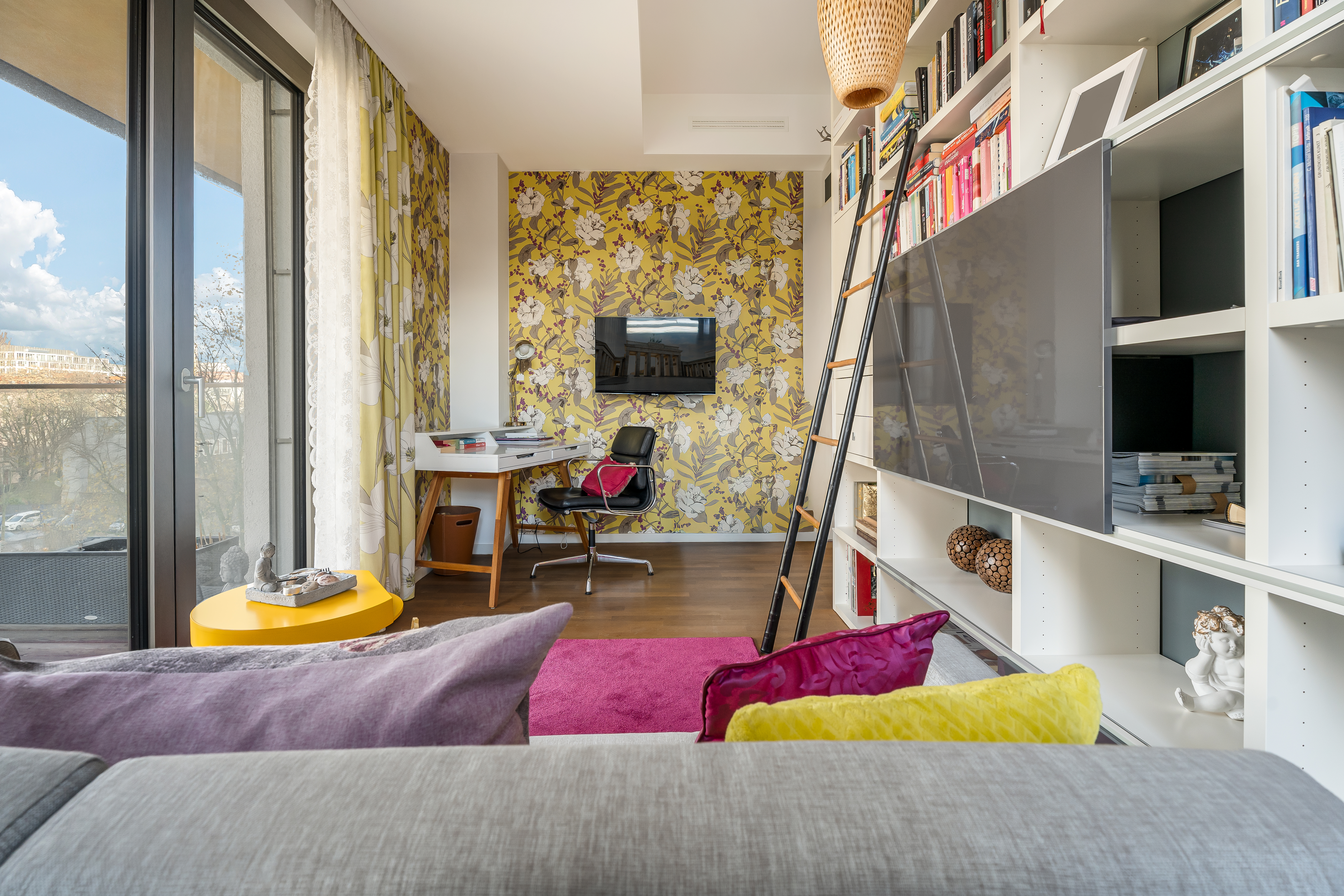 Modernes Wohnzimmer mit bunten Kissen, großem Regal, Schreibtisch und gelbem Beistelltisch am Fenster.