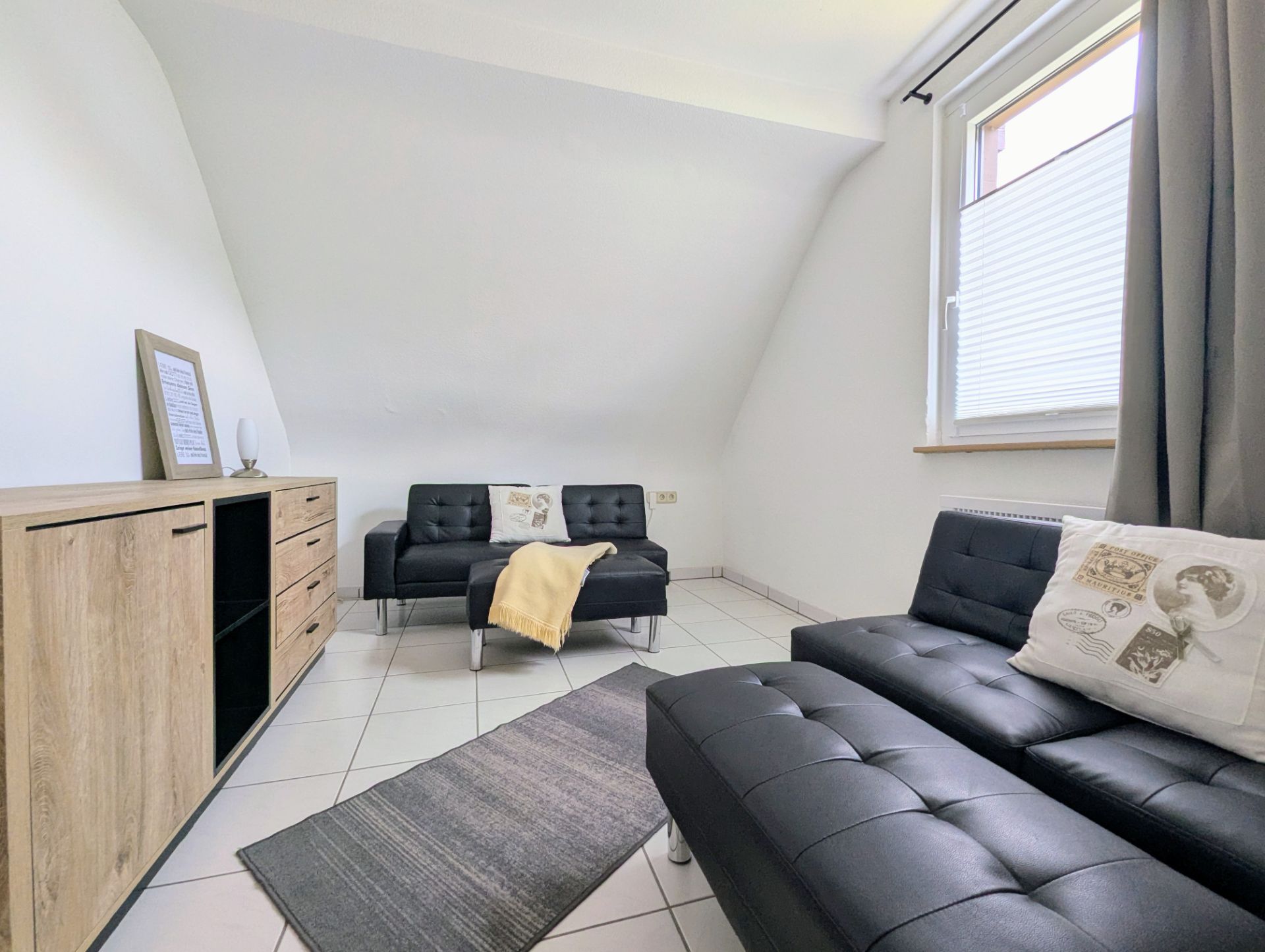 Wohnzimmer mit zwei schwarzen Sofas, Teppich, Holzkommode und Fenster mit grauem Vorhang.