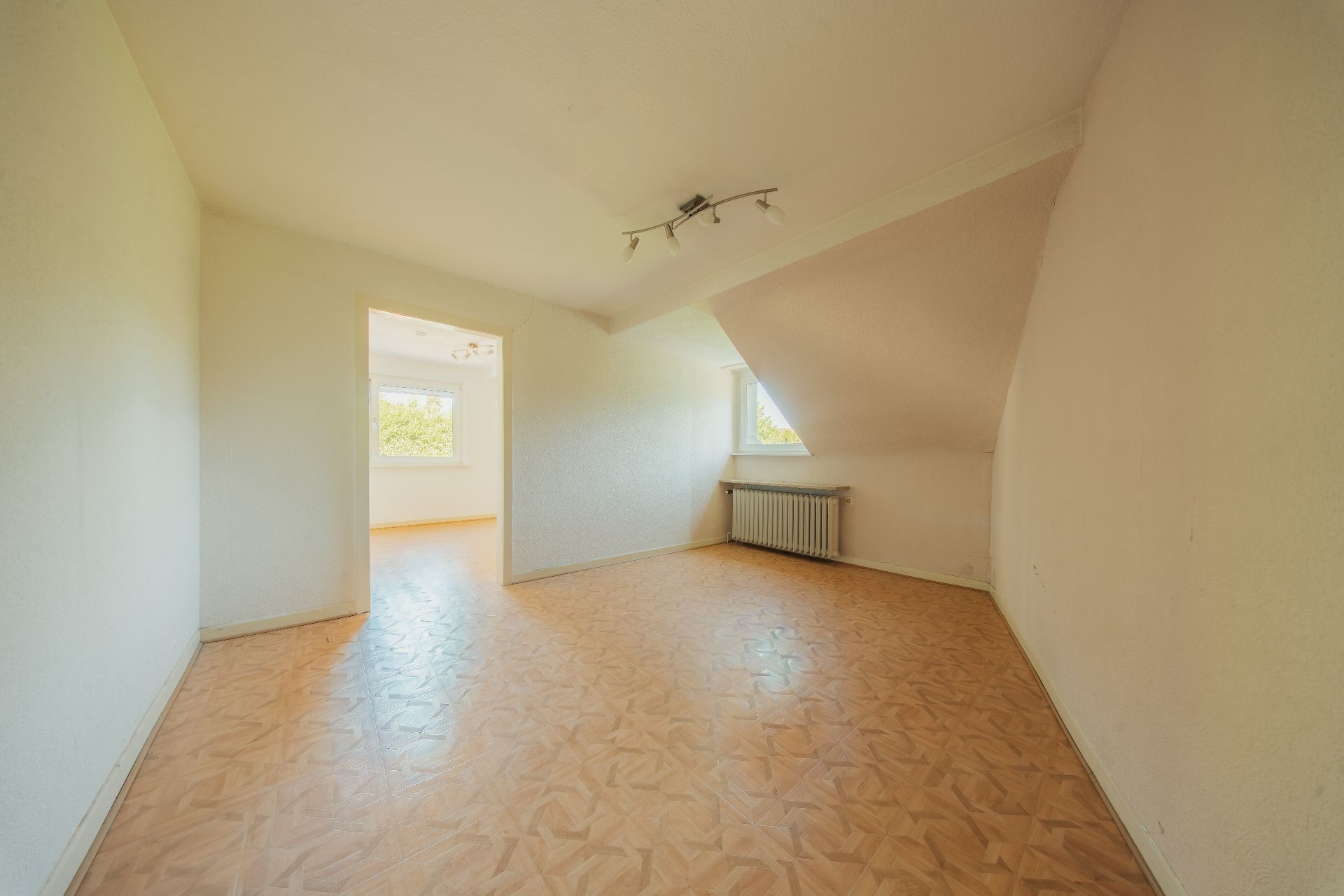 Leerer Raum mit schräger Decke, Holzboden, Fenster und Heizung, angrenzender Raum sichtbar.