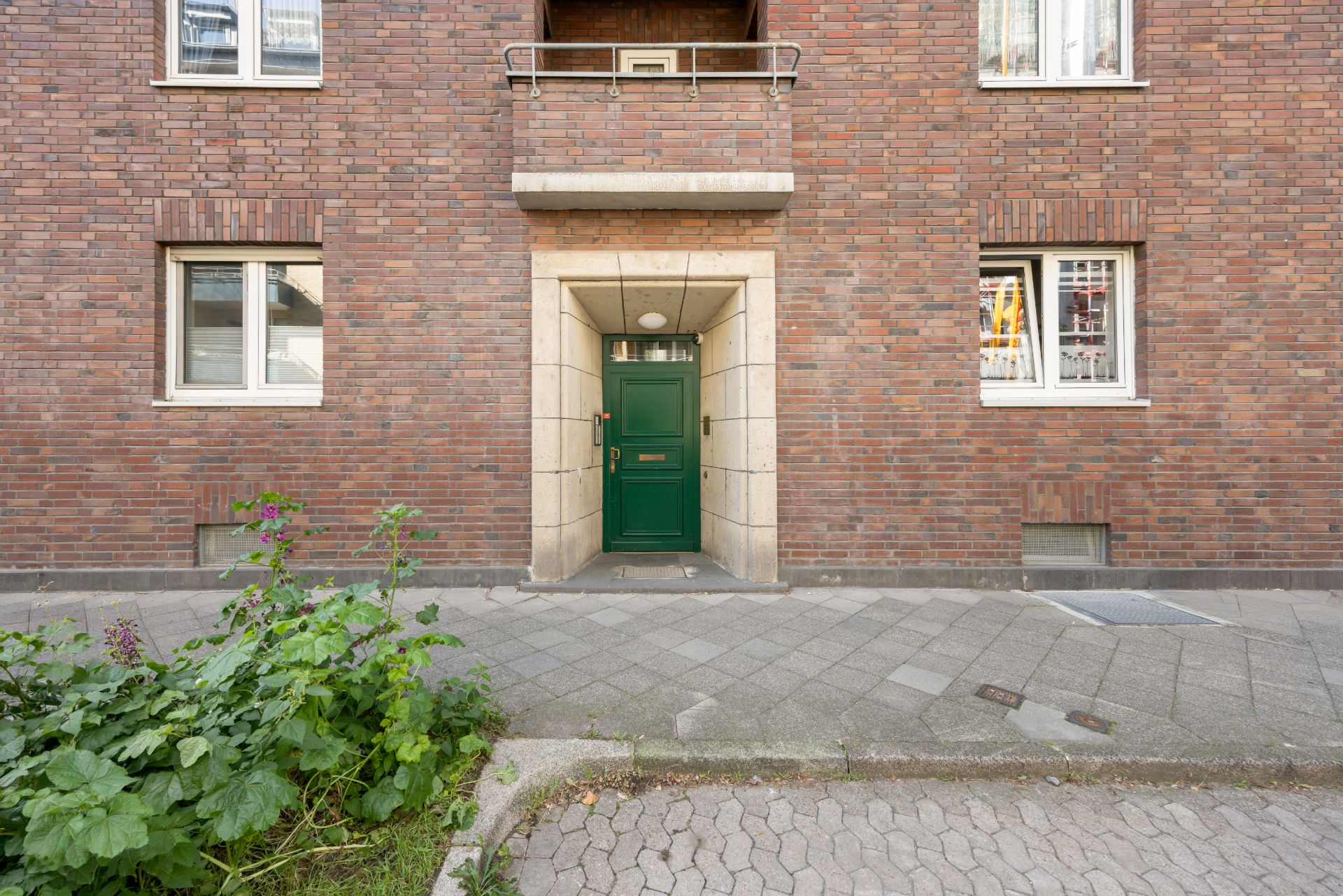 Grüne Haustür in einer Backsteinfassade mit zwei Fenstern und kleinen Pflanzen davor.