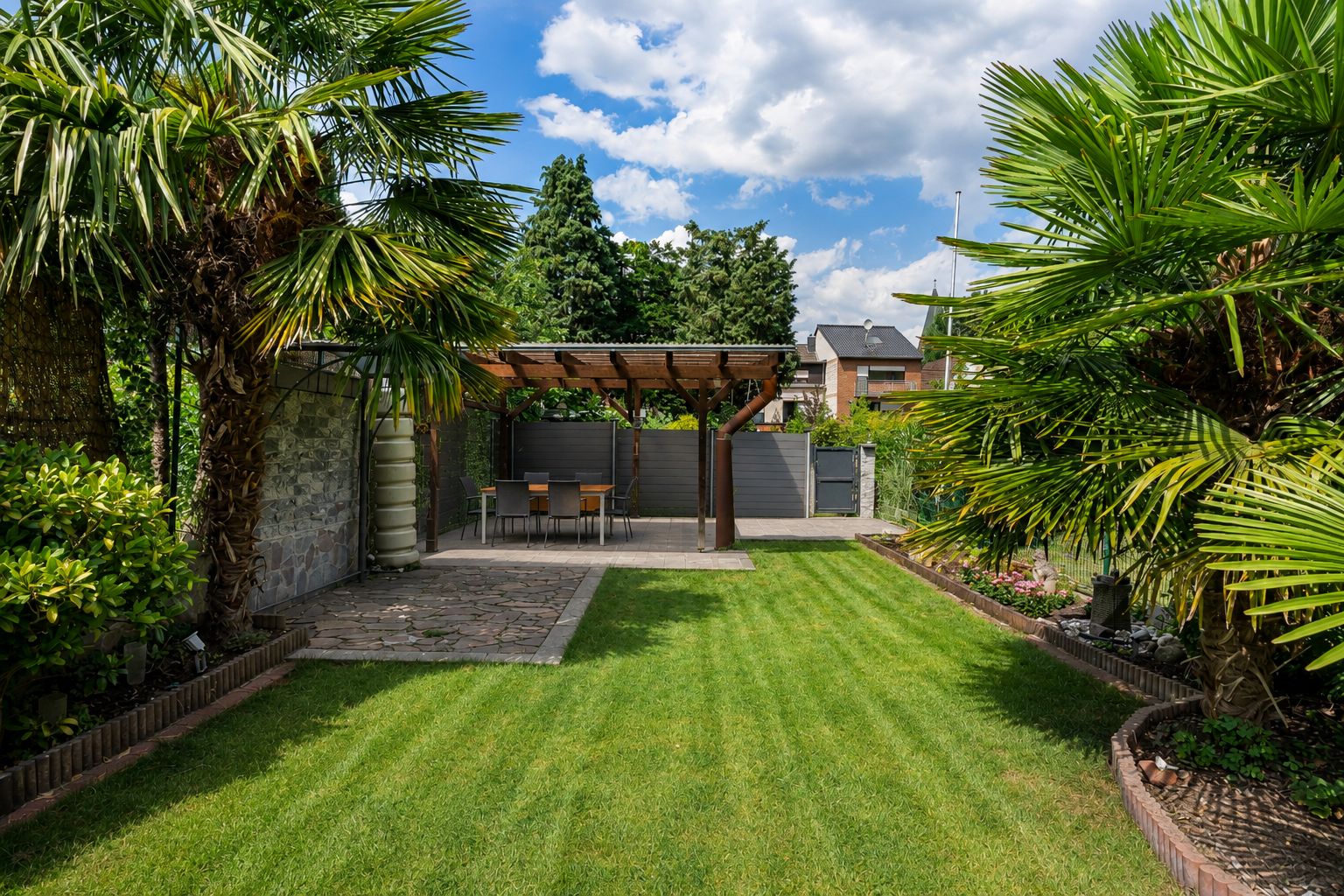 Garten mit Rasenfläche, Palmen und überdachter Terrasse mit Tisch und Stühlen.
