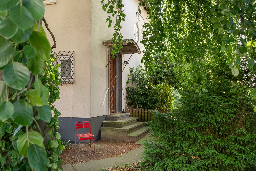 Eingang eines Wohnhauses mit rotem Stuhl und viel grüner Bepflanzung im Vordergrund.