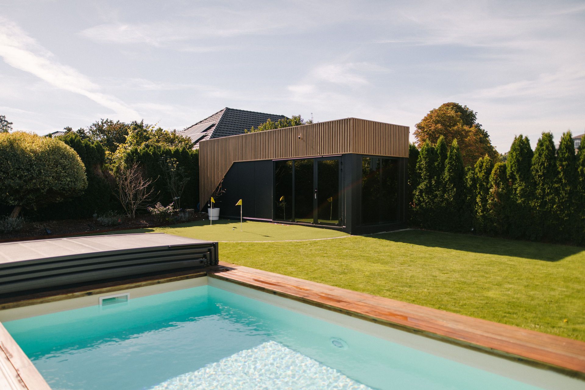 Garten mit modernem Gebäude, Pool und Rasenfläche, umgeben von Bäumen und Sträuchern.