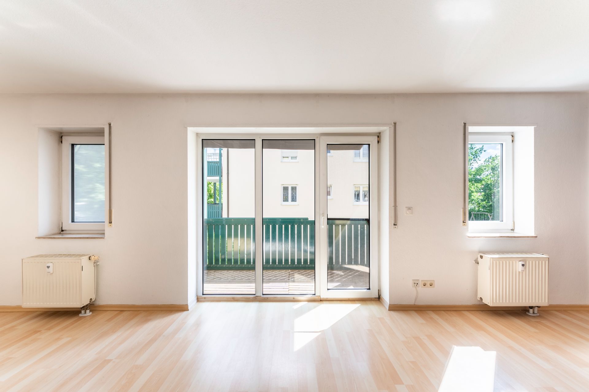 Helles Zimmer mit Parkettboden, zwei Fenstern und einer Glastür zu einem Balkon.