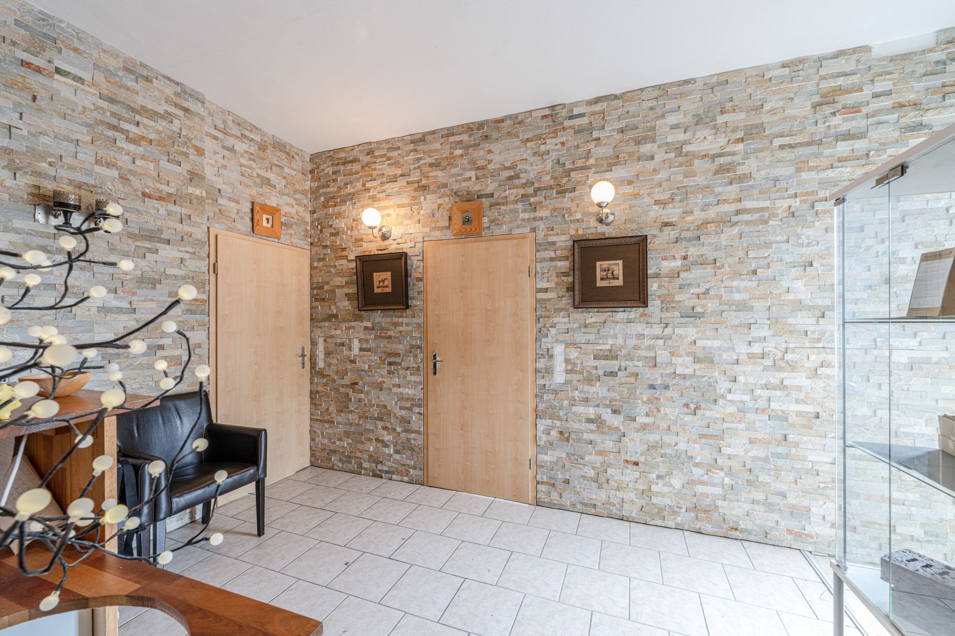 Flur mit zwei Holztüren, Steinwand, schwarzen Sesseln und Wandbildern.