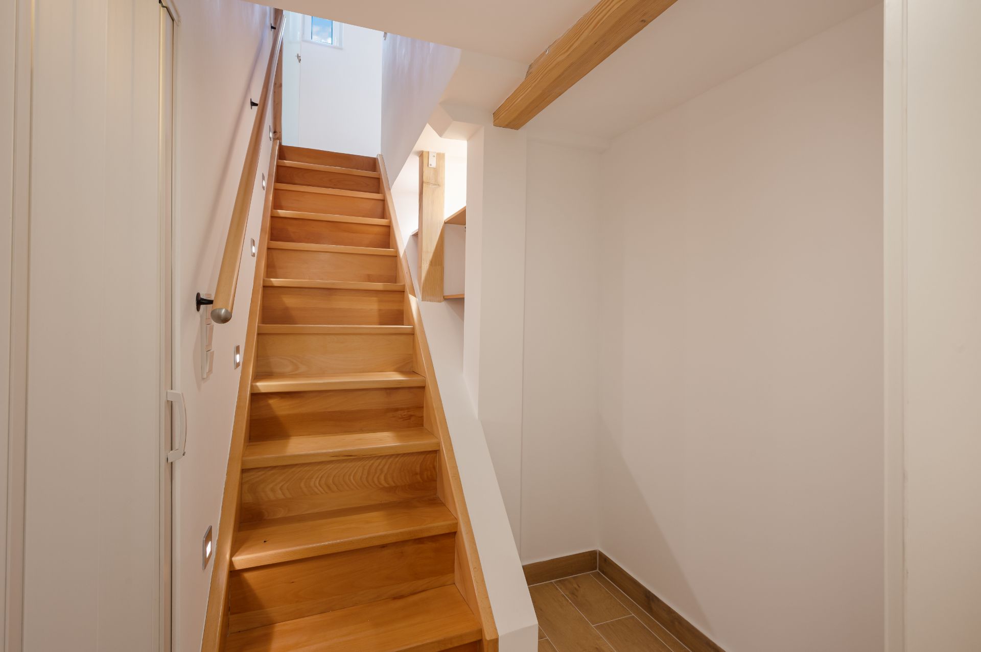 Holztreppe mit Handlauf in einem hellen Flur, oben ein kleines Fenster.