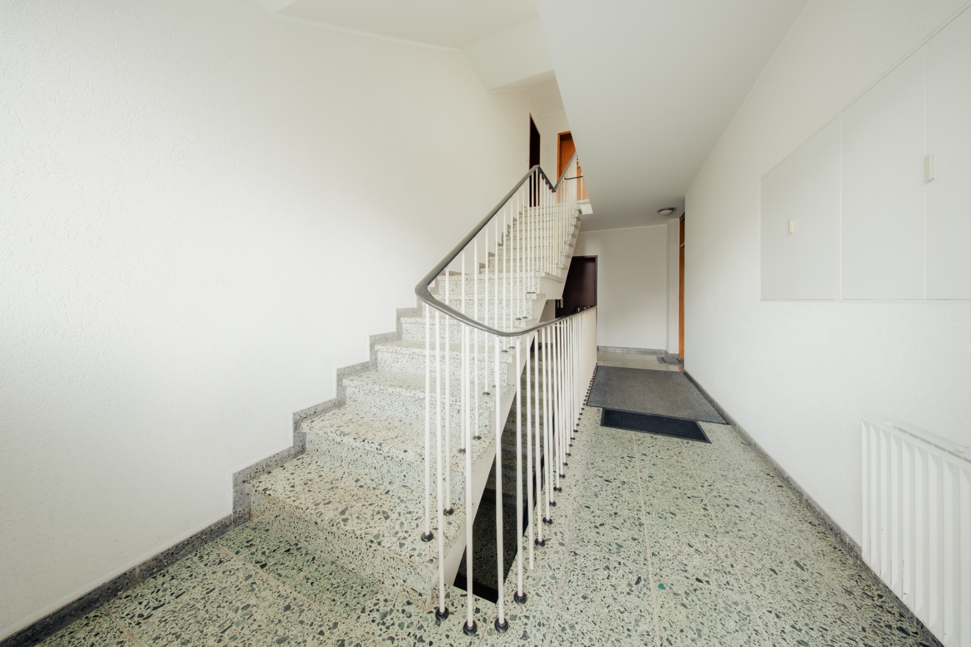 Heller Hausflur mit Treppe und Handlauf, gefliester Boden und weißen Wänden.