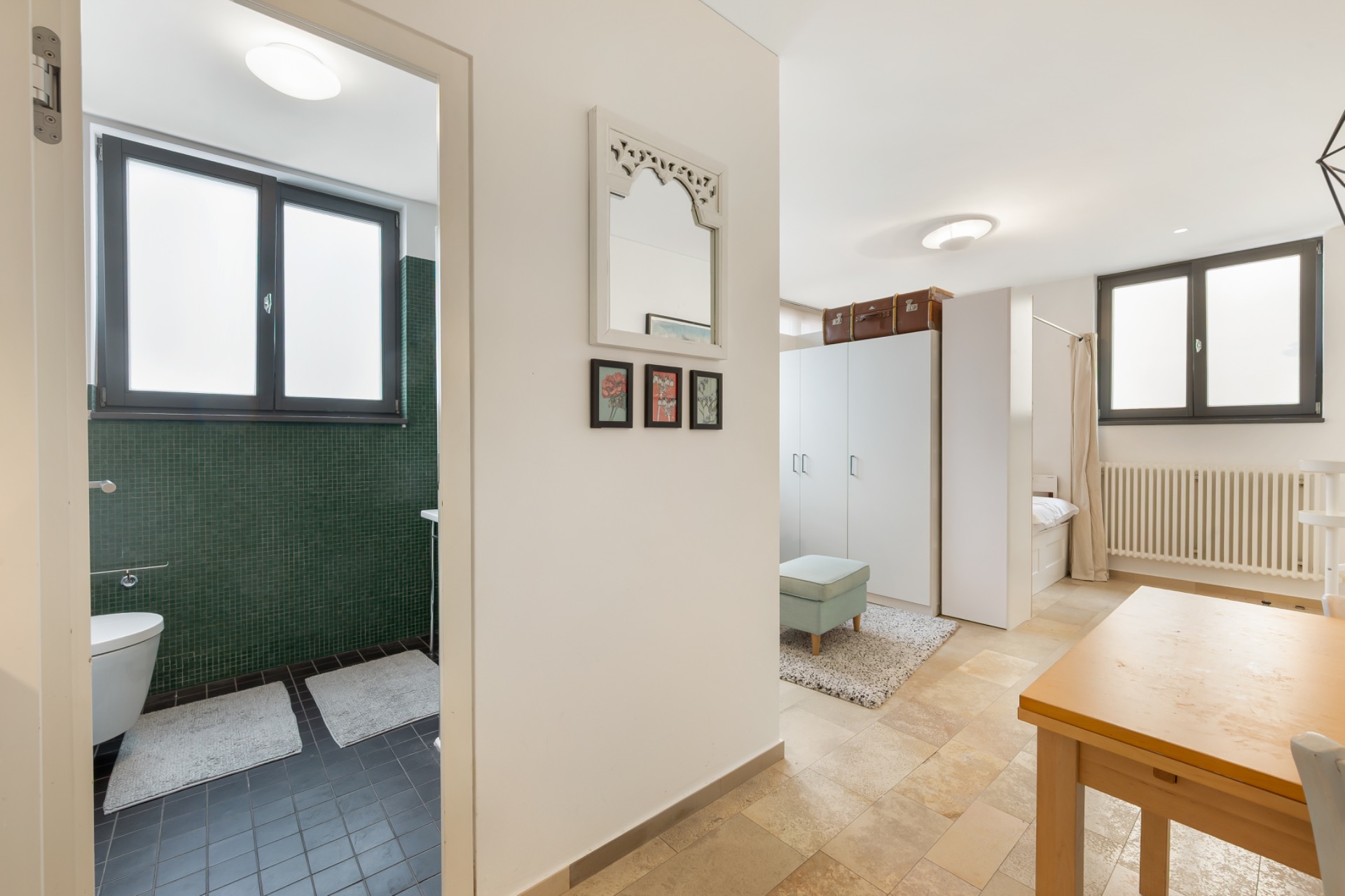 Heller Wohnraum mit Blick ins Bad, zwei Fenstern, Schrank und Holztisch.
