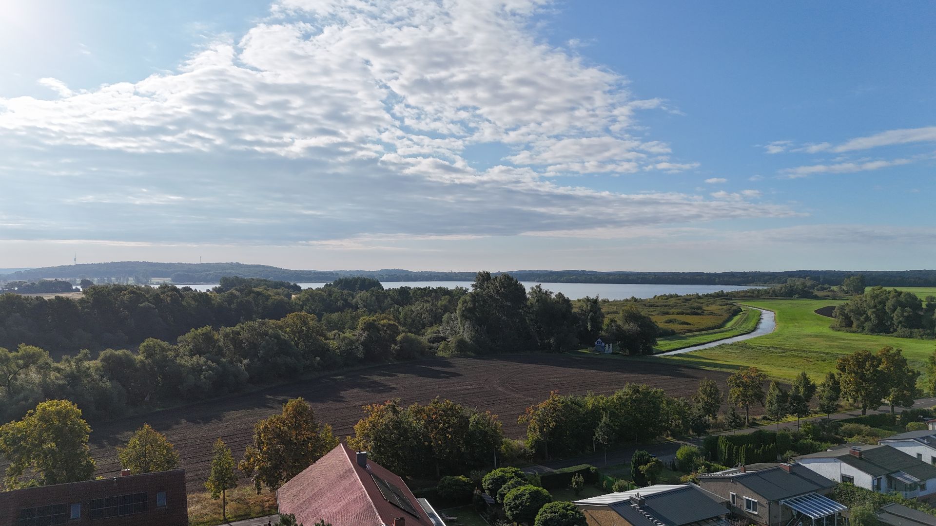 Landschaft mit Häusern im Vordergrund, Feldern, Bäumen und einem Fluss unter teils bewölktem Himmel.