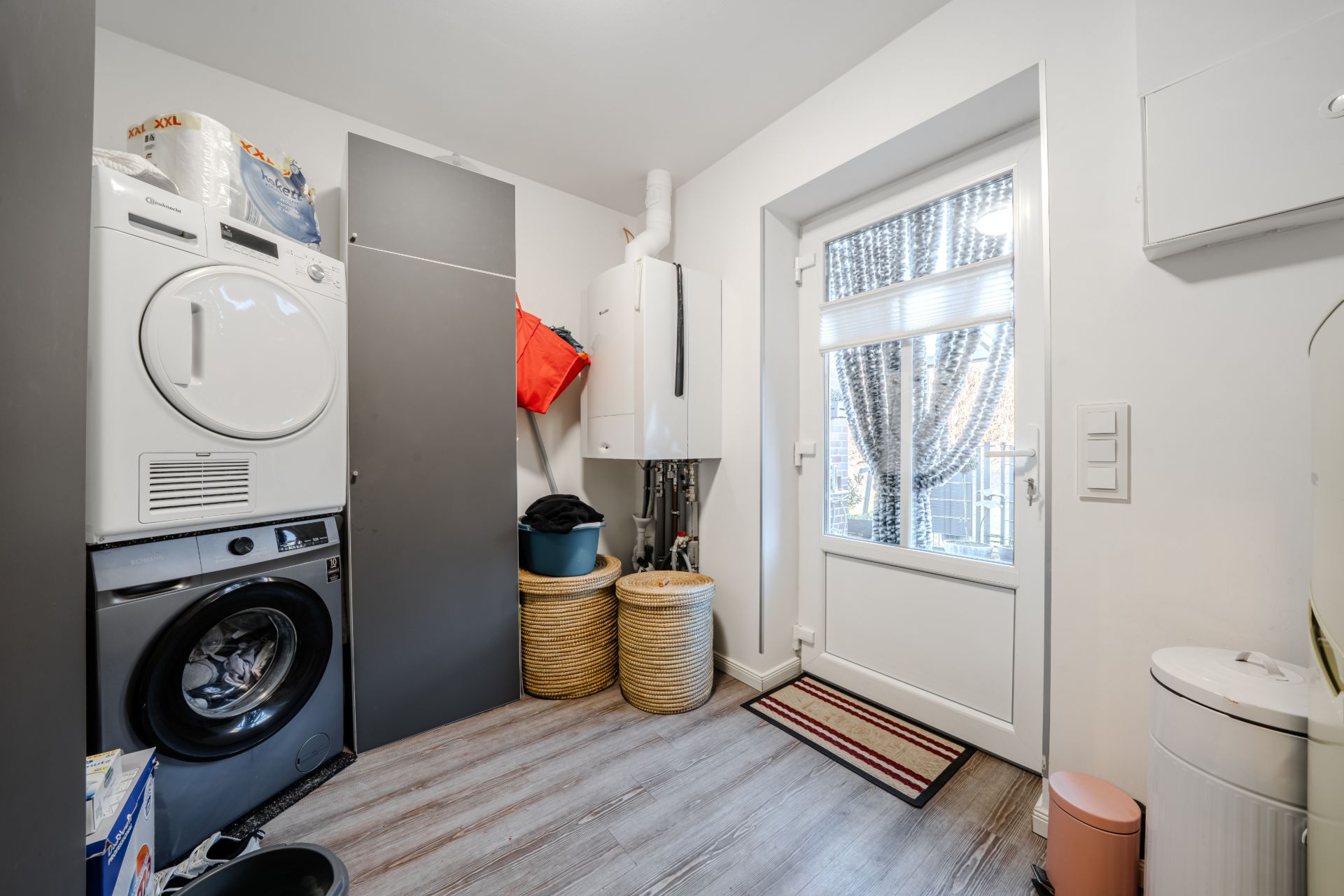 Hauswirtschaftsraum mit Waschmaschine, Trockner, Schränken, Wäschekörben und Tür mit Fenstervorhang.