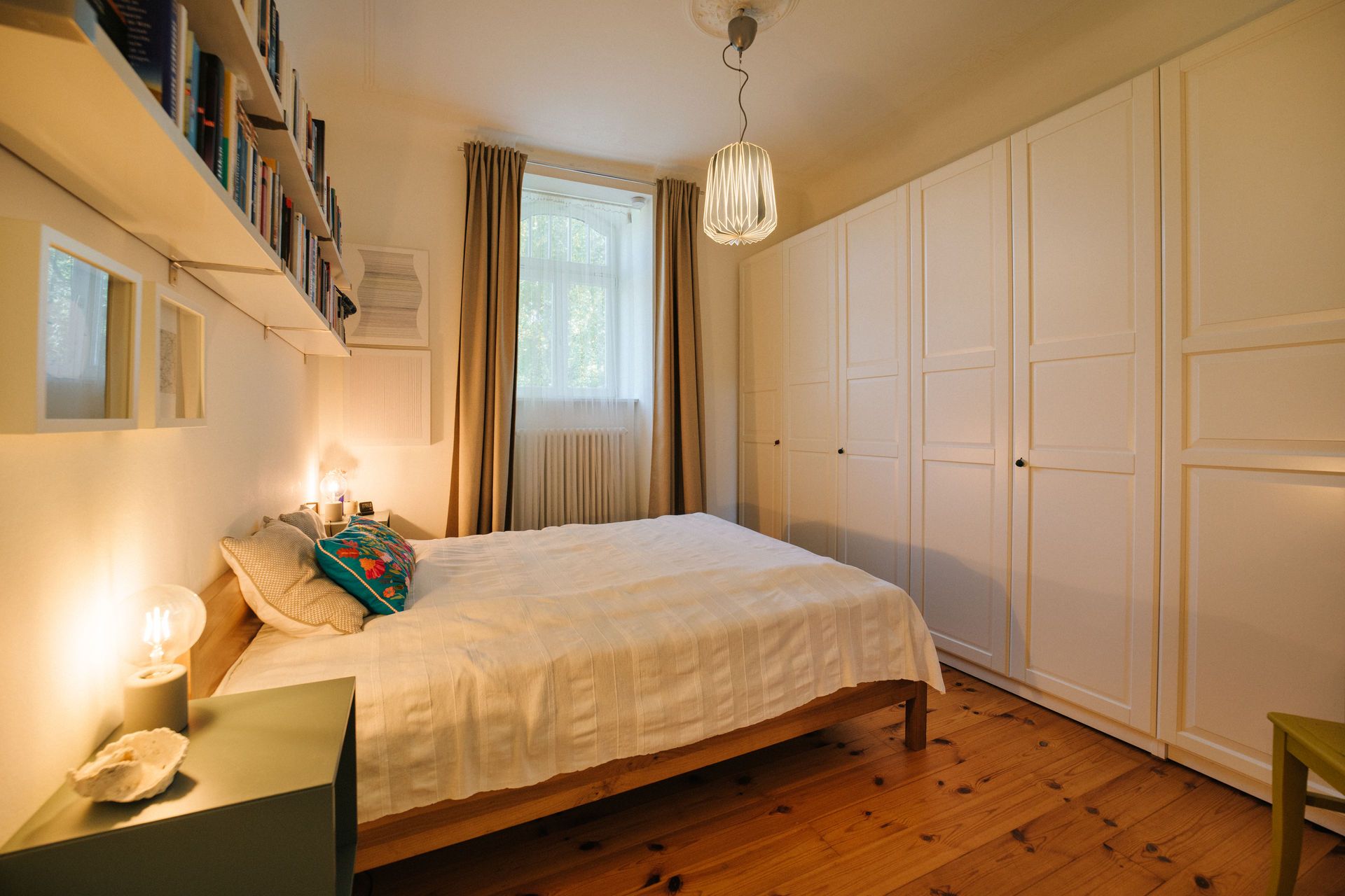 Helles Schlafzimmer mit Doppelbett, Bücherregal, großem Kleiderschrank und Fenster mit Vorhängen.