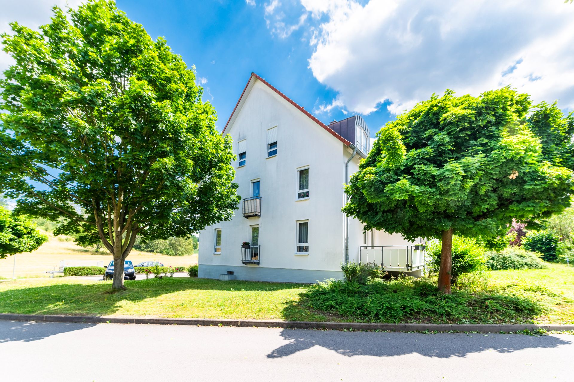 Weißes Wohnhaus mit mehreren Fenstern, umgeben von grünen Bäumen und Rasen.
