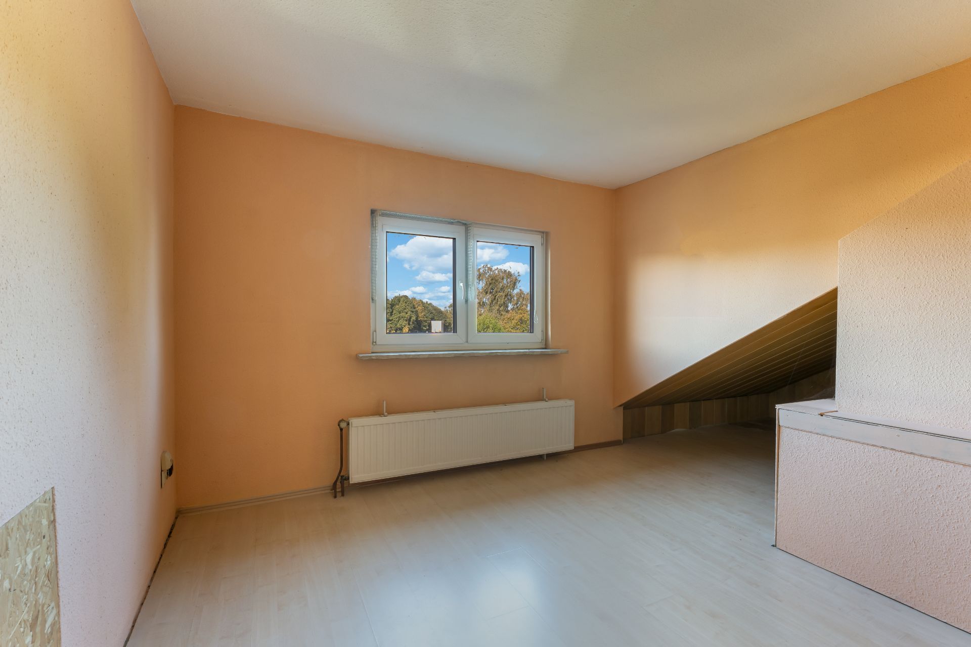 Leerer Raum mit Dachschräge, Fenster und Heizkörper, Wände in Orange gestrichen.