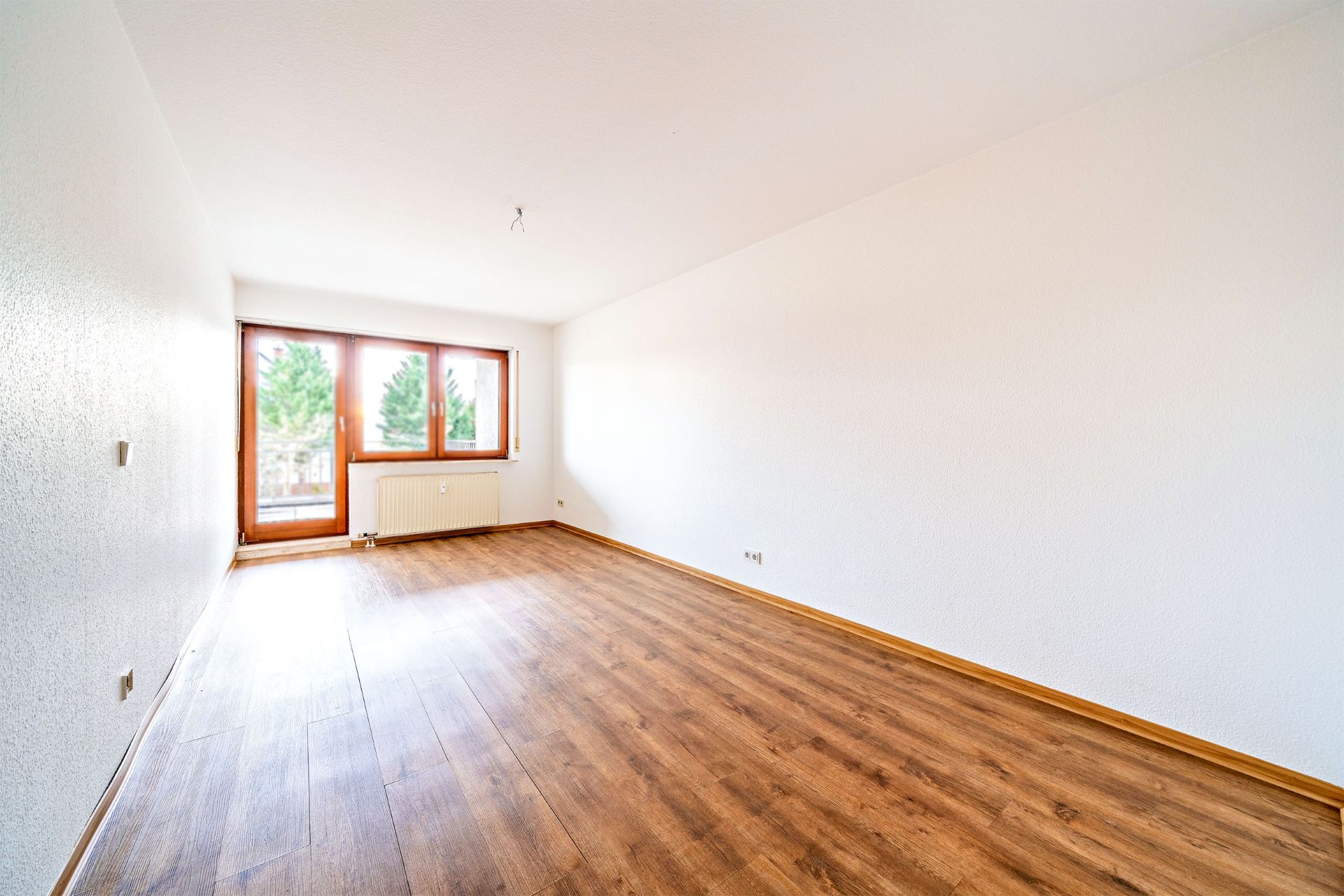 Leerer Raum mit weißer Wand, Holzboden und großem Fenster am Ende.