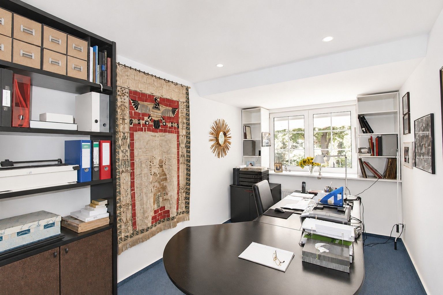 Helles Arbeitszimmer mit großem Schreibtisch, Regalen, Wandteppich und Fenster mit Blick ins Grüne.