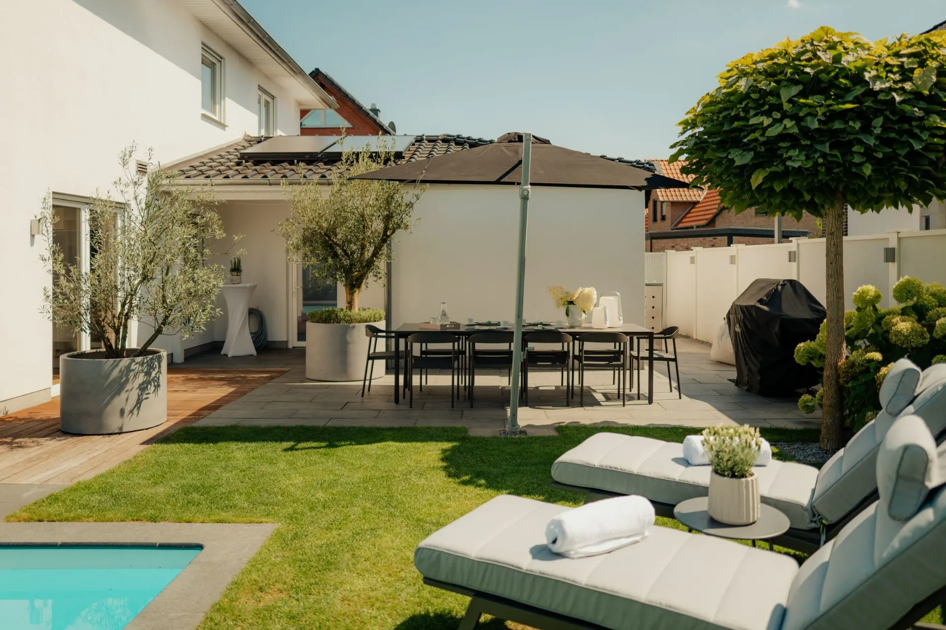Garten mit Liegen, Esstisch, Sonnenschirm, Grill und Pool neben einem modernen Wohnhaus.