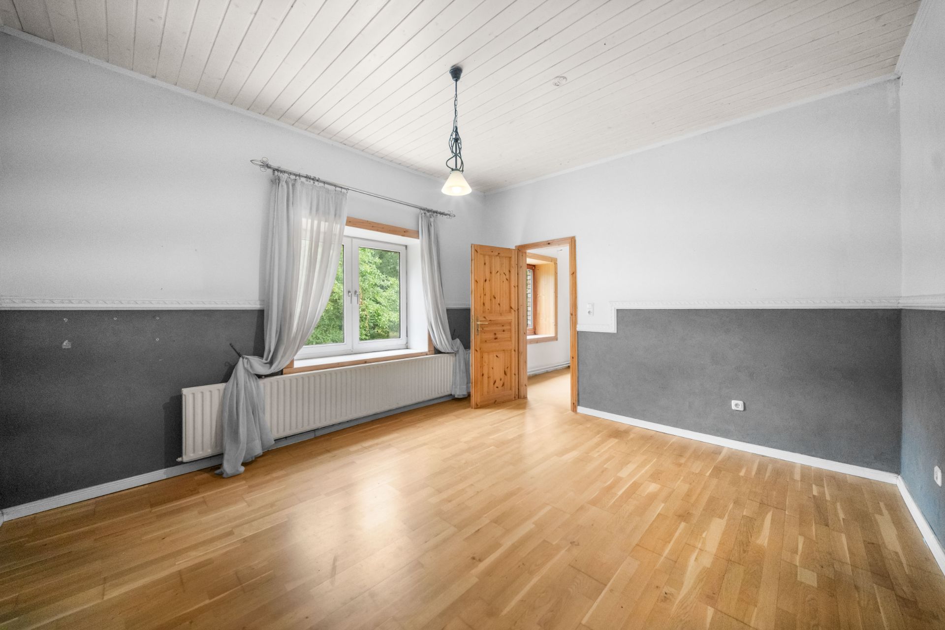 Helles Zimmer mit Holzboden, einem Fenster mit Vorhängen und einer offenen Holztür.