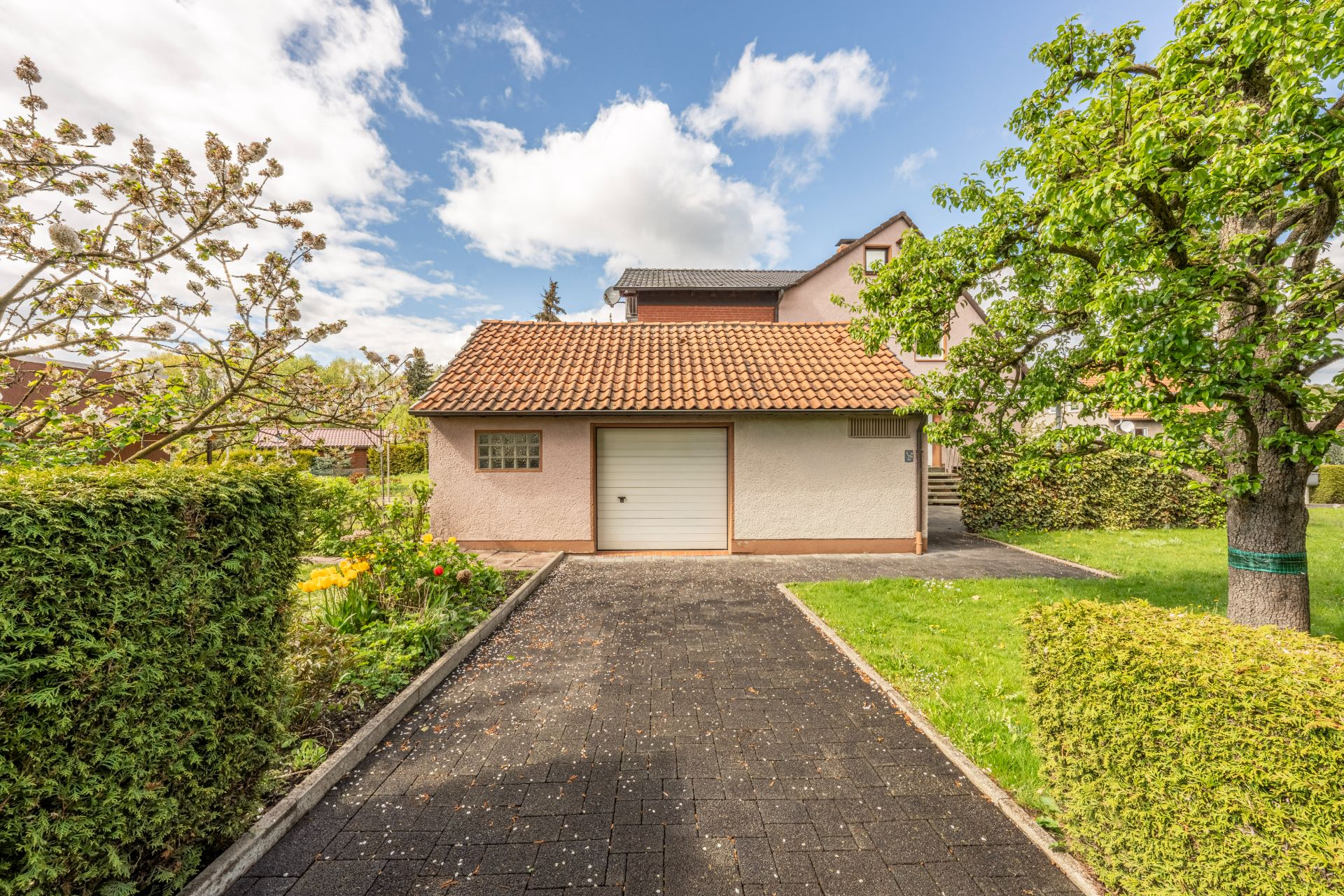 Garage mit weißem Tor, gepflasterte Einfahrt, Garten und Baum im Vordergrund.