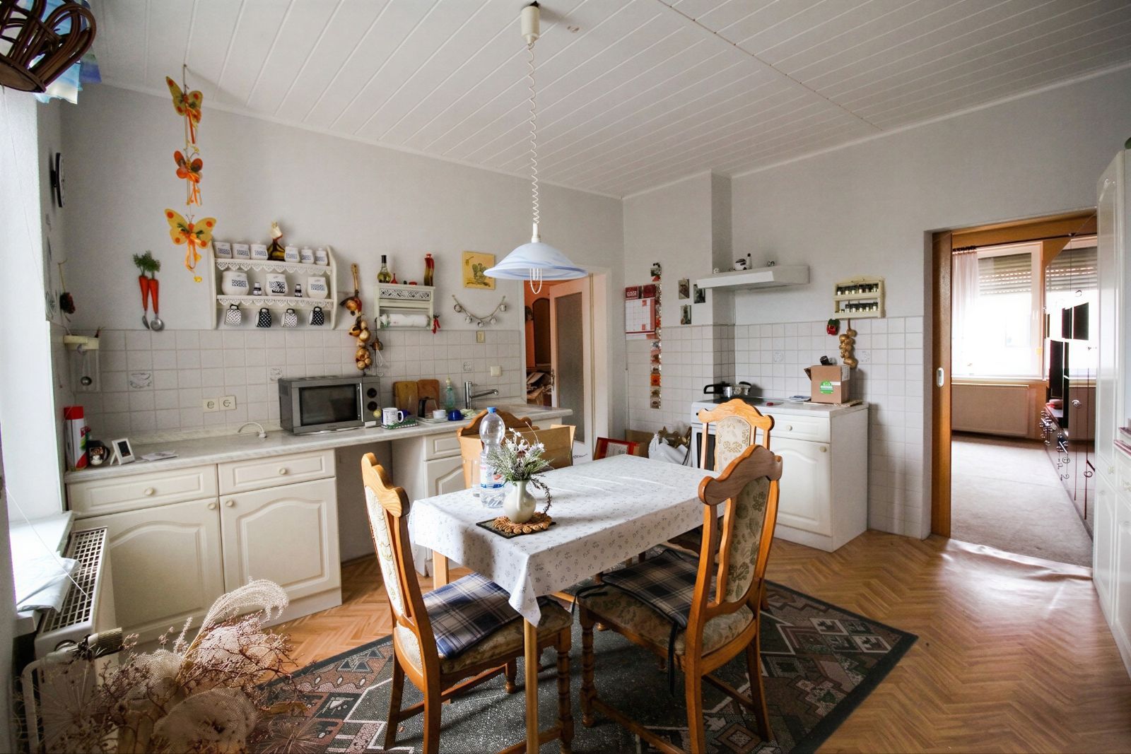 Küche mit Esstisch, vier Stühlen, Hängelampe und offener Tür zum angrenzenden Zimmer.