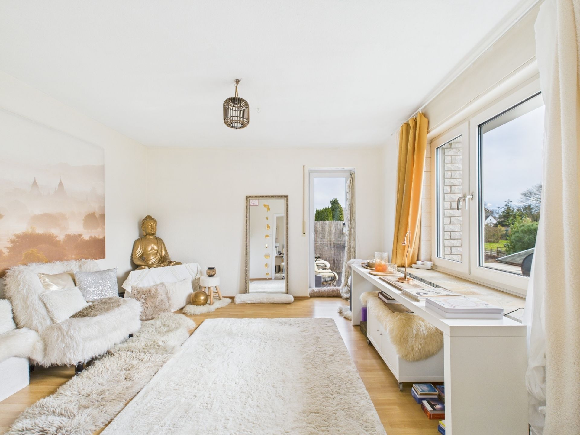Helles Wohnzimmer mit Sofa, Teppichen, Buddha-Figur, Spiegel und großem Fenster.