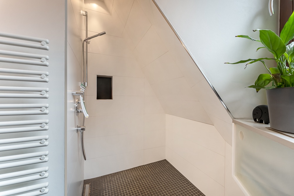 Moderne Dusche unter Dachschräge mit weißem Heizkörper und Pflanze auf Fensterbank.