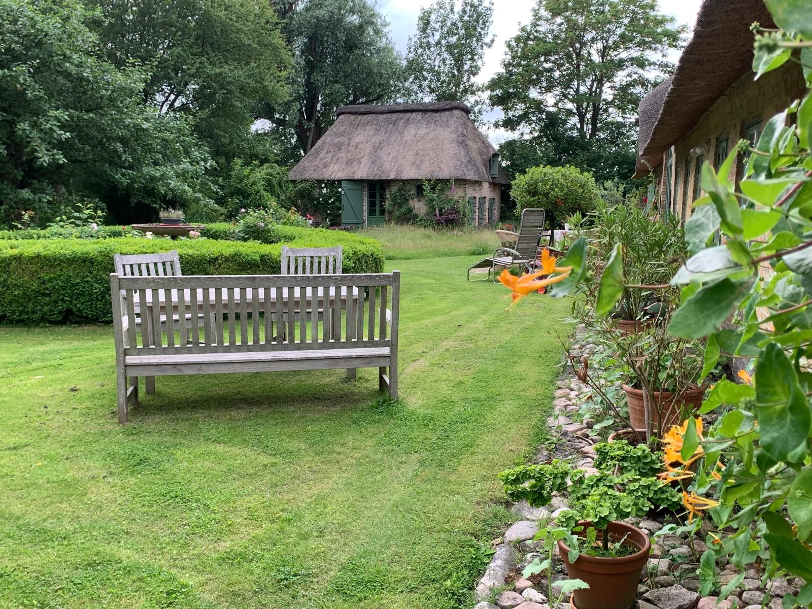 Grüner Garten mit Holzbänken, Blumentöpfen und Haus mit Reetdach im Hintergrund.
