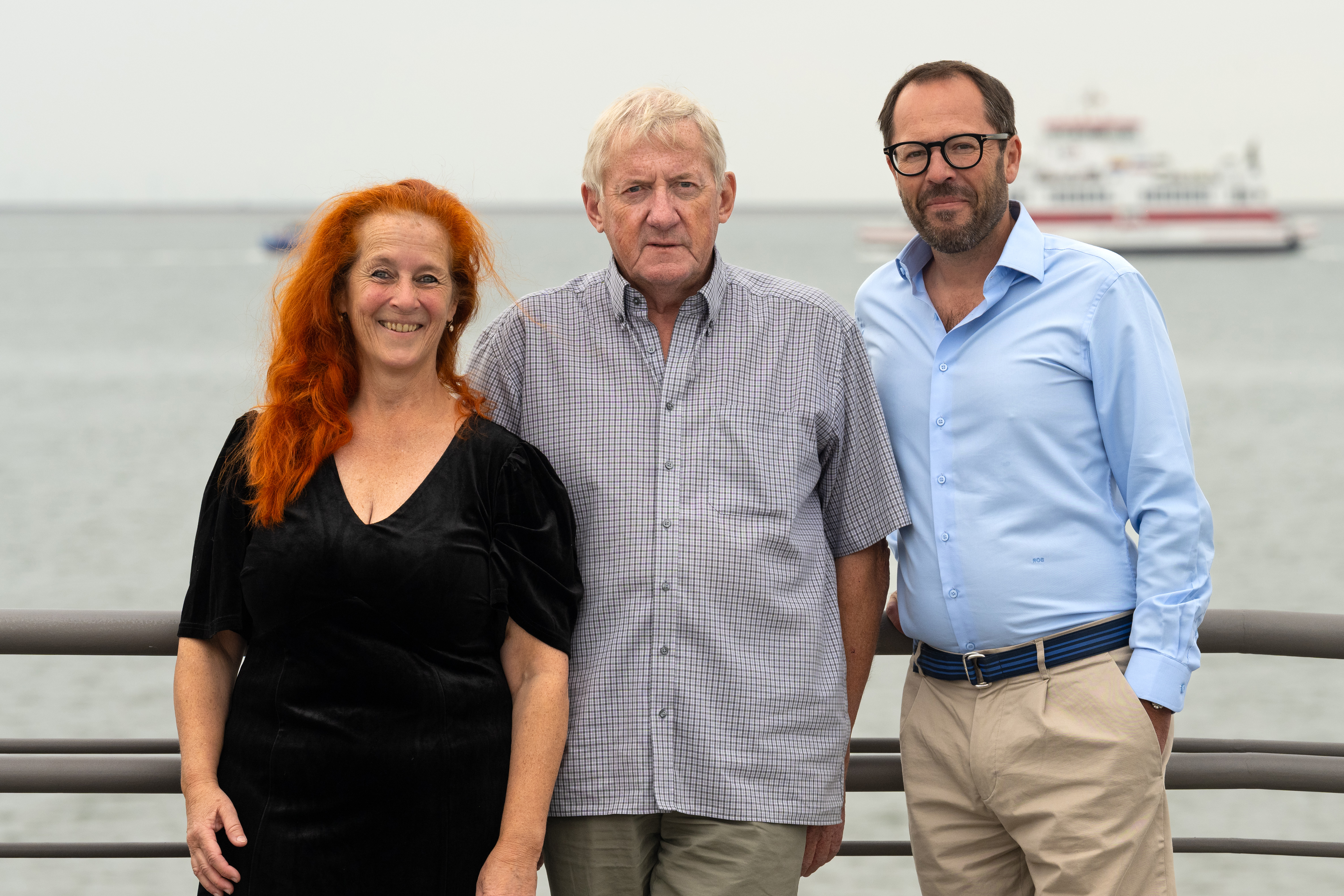 Drei Personen stehen an einem Geländer vor Wasser, im Hintergrund ist ein Schiff zu sehen.