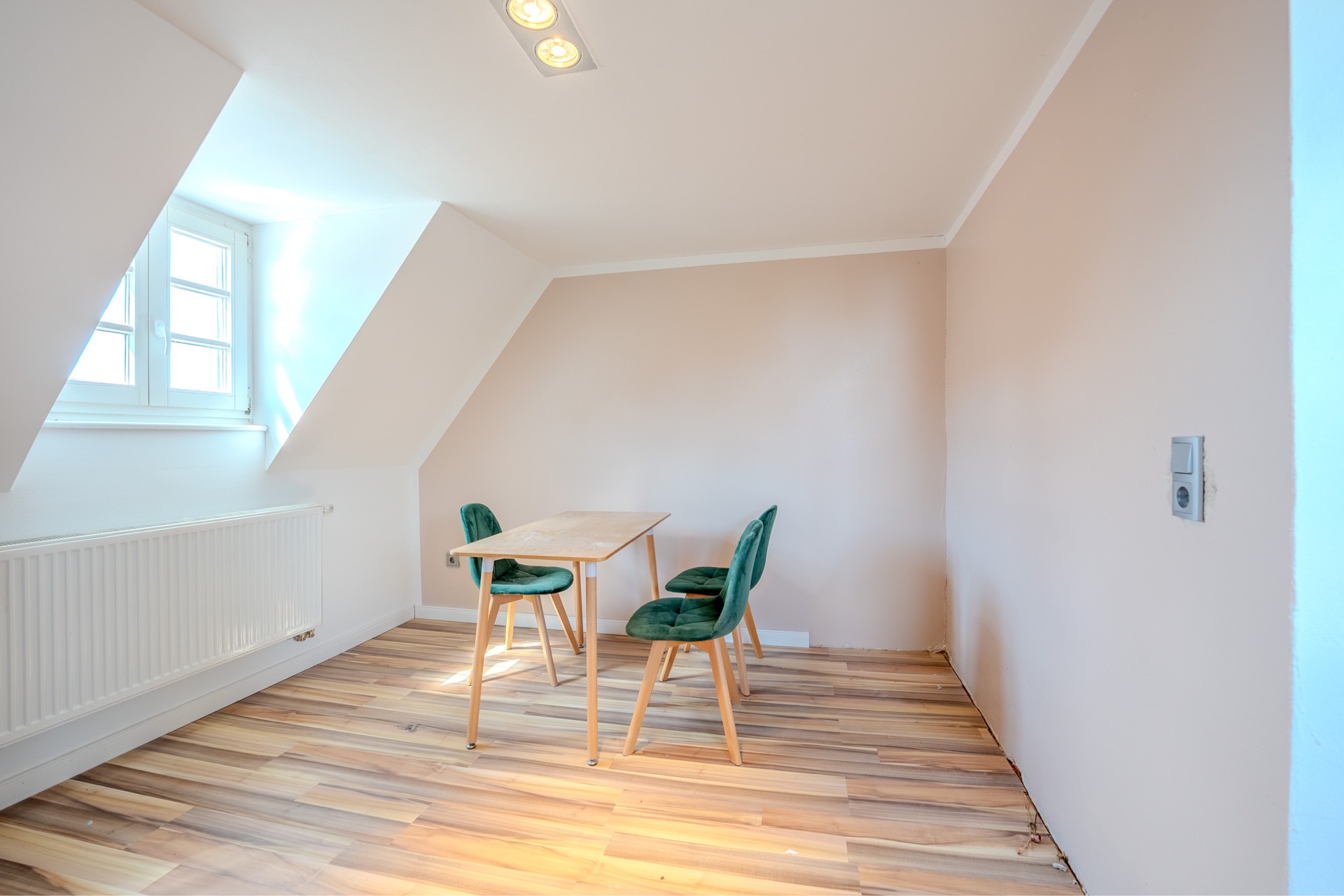 Kleiner Raum mit Holzfußboden, Tisch und drei grünen Stühlen, Fenster an der linken Wand.