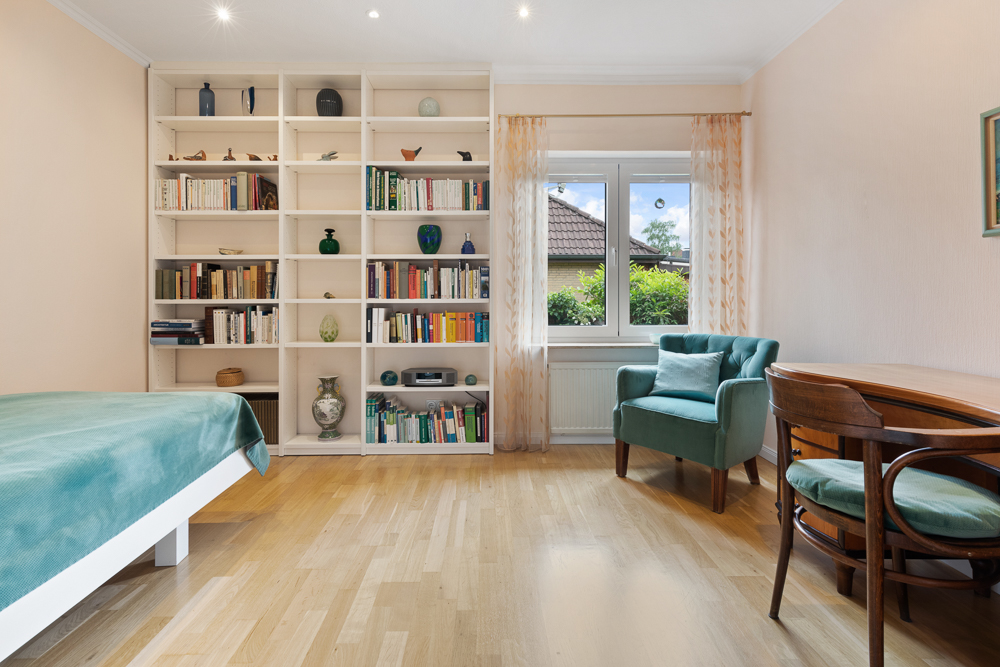 Helles Zimmer mit Bücherregal, Sessel, Schreibtisch und Bett mit grüner Decke.