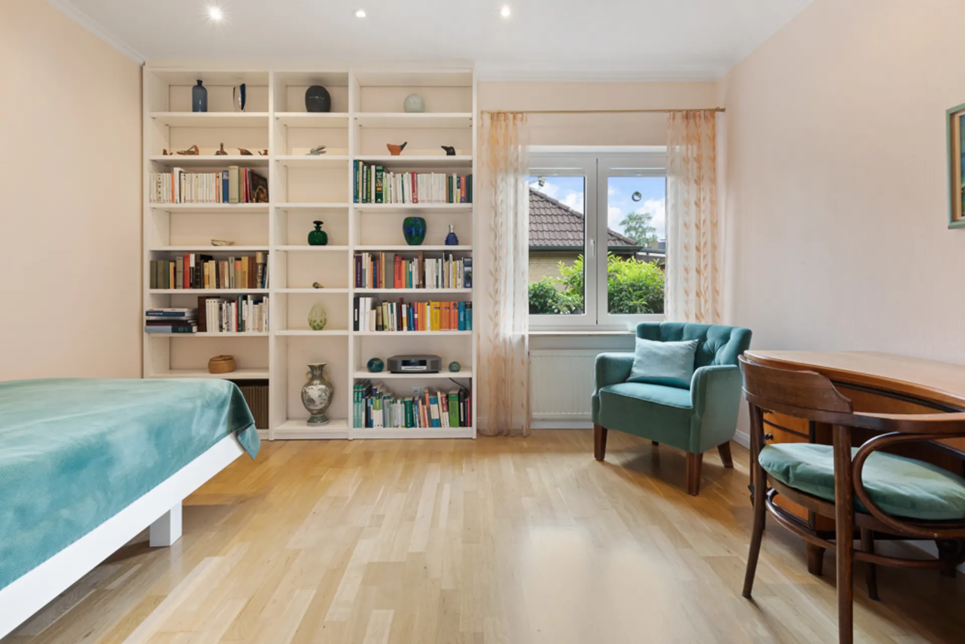 Helles Zimmer mit Bücherregal, Sessel, Schreibtisch und Bett mit grüner Decke.