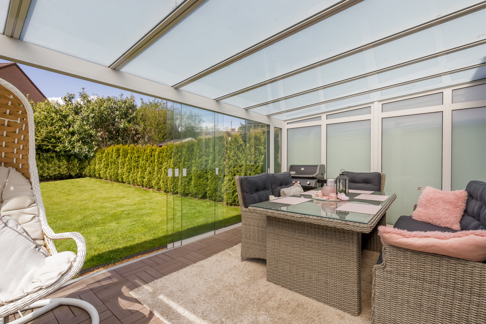 Überdachte Terrasse mit Glaswänden, Sitzgruppe, Grill und Blick auf einen grünen Garten.