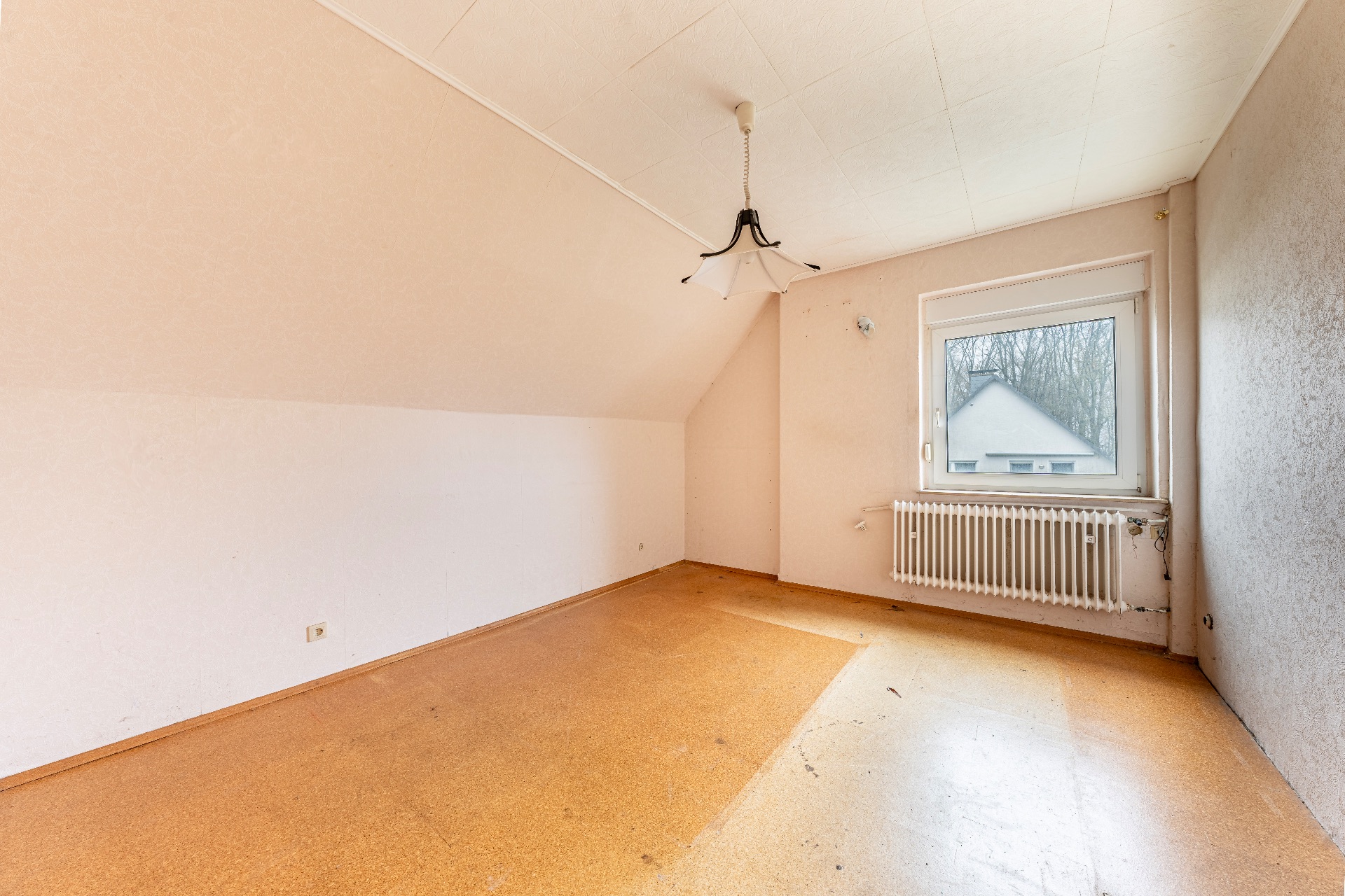 Leeres Zimmer mit schräger Decke, Fenster, Heizkörper und einfacher Deckenlampe.