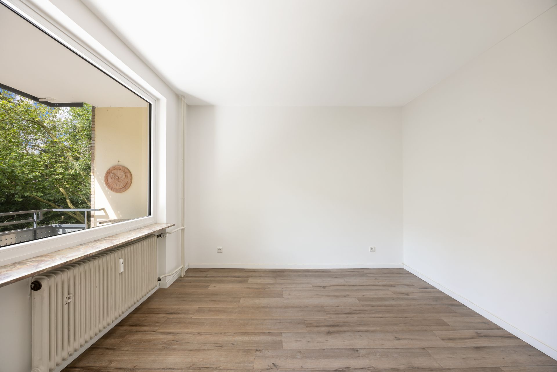Helles, leeres Zimmer mit großem Fenster und Holzfußboden.