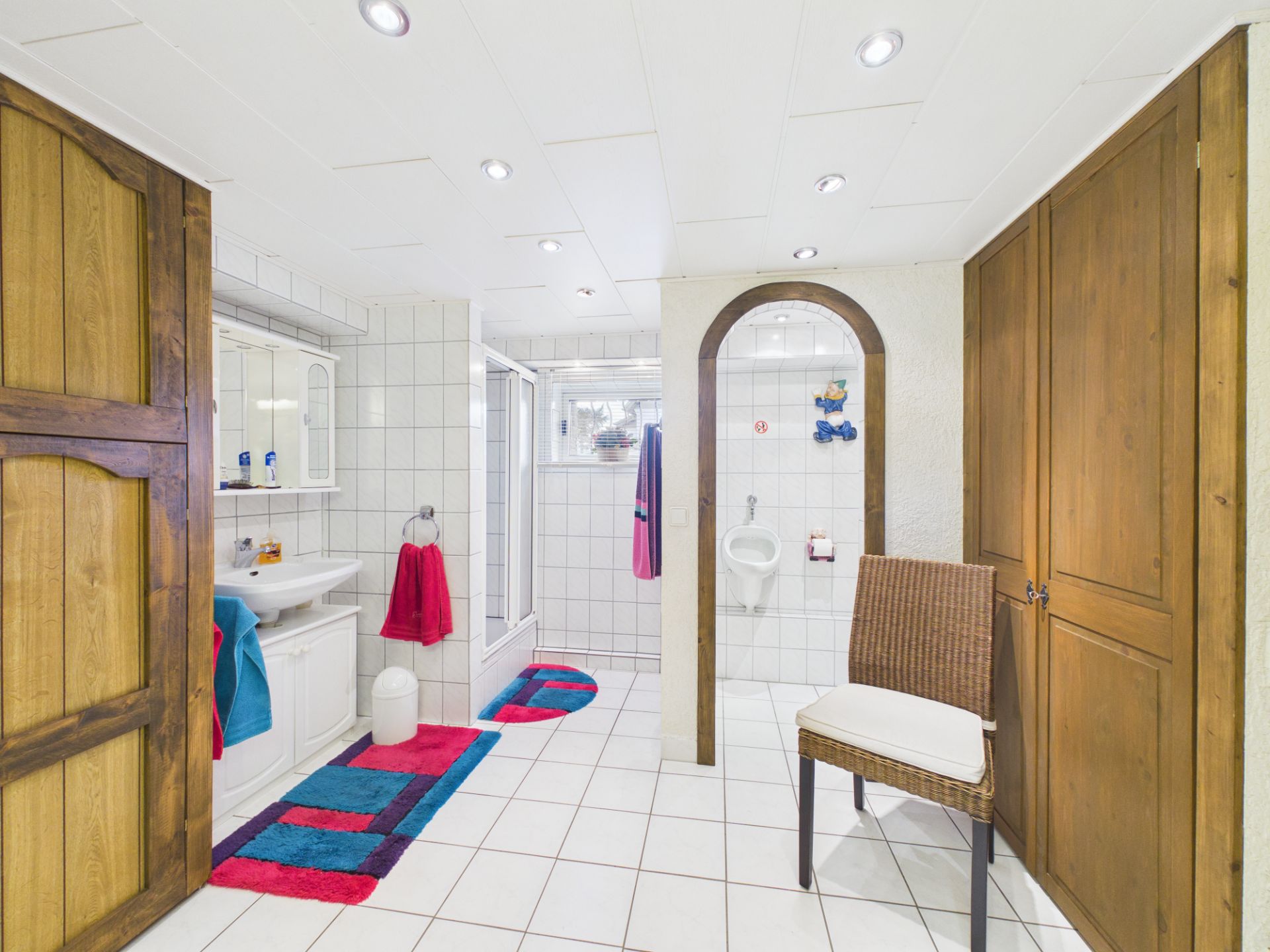 Helles Badezimmer mit weißen Fliesen, Waschbecken, Dusche, WC und bunten Teppichen.