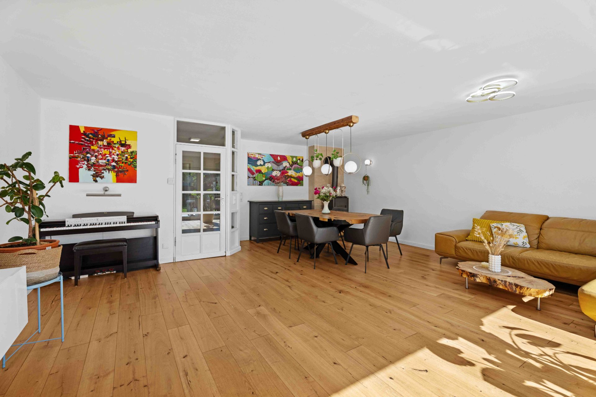 Helles Wohnzimmer mit Holzboden, Esstisch, Sofa, Klavier und bunten Bildern an der Wand.