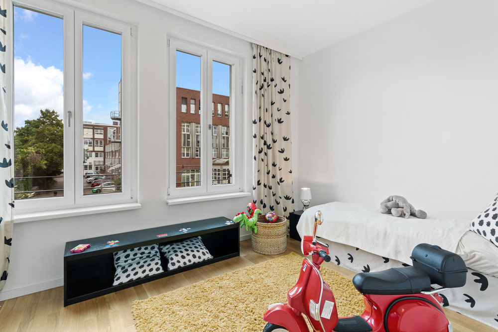 Helles Kinderzimmer mit Bett, Spielzeug-Roller, Teppich und großen Fenstern.