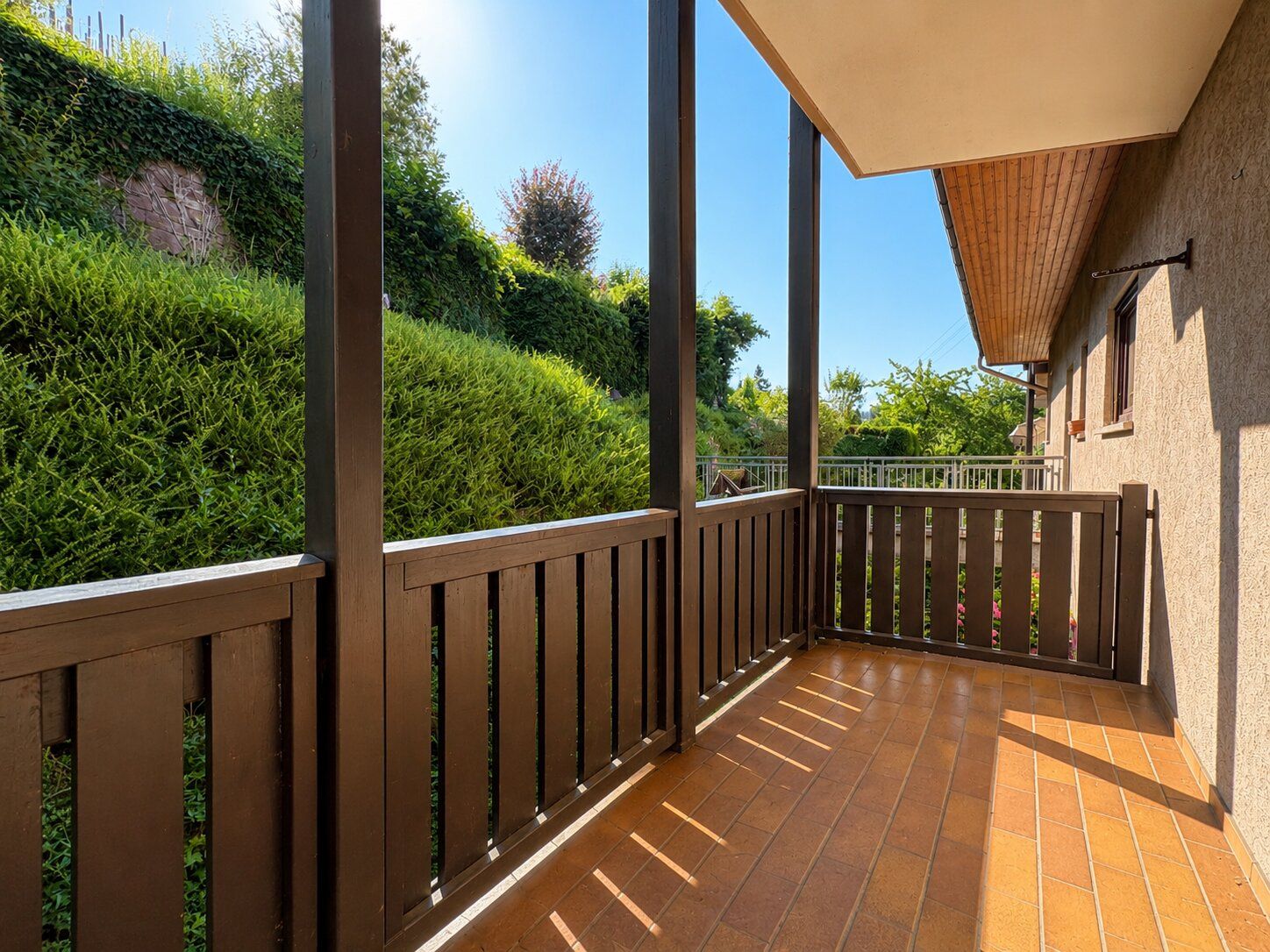 Überdachte Terrasse mit braunem Geländer und Blick auf einen grünen Garten.