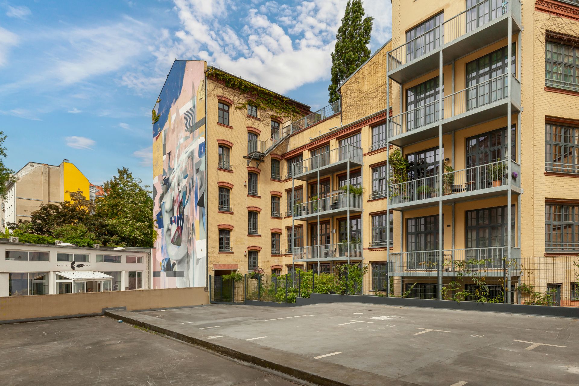 Innenhof mit gelbem Wohngebäude und Balkonen, Parkfläche im Vordergrund, Wandbild links.