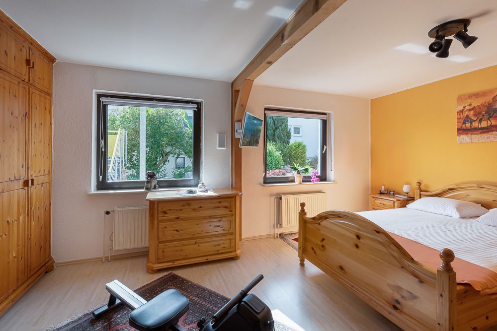 Helles Schlafzimmer mit Holzmöbeln, zwei Fenstern und einem Fitnessgerät auf Teppich.