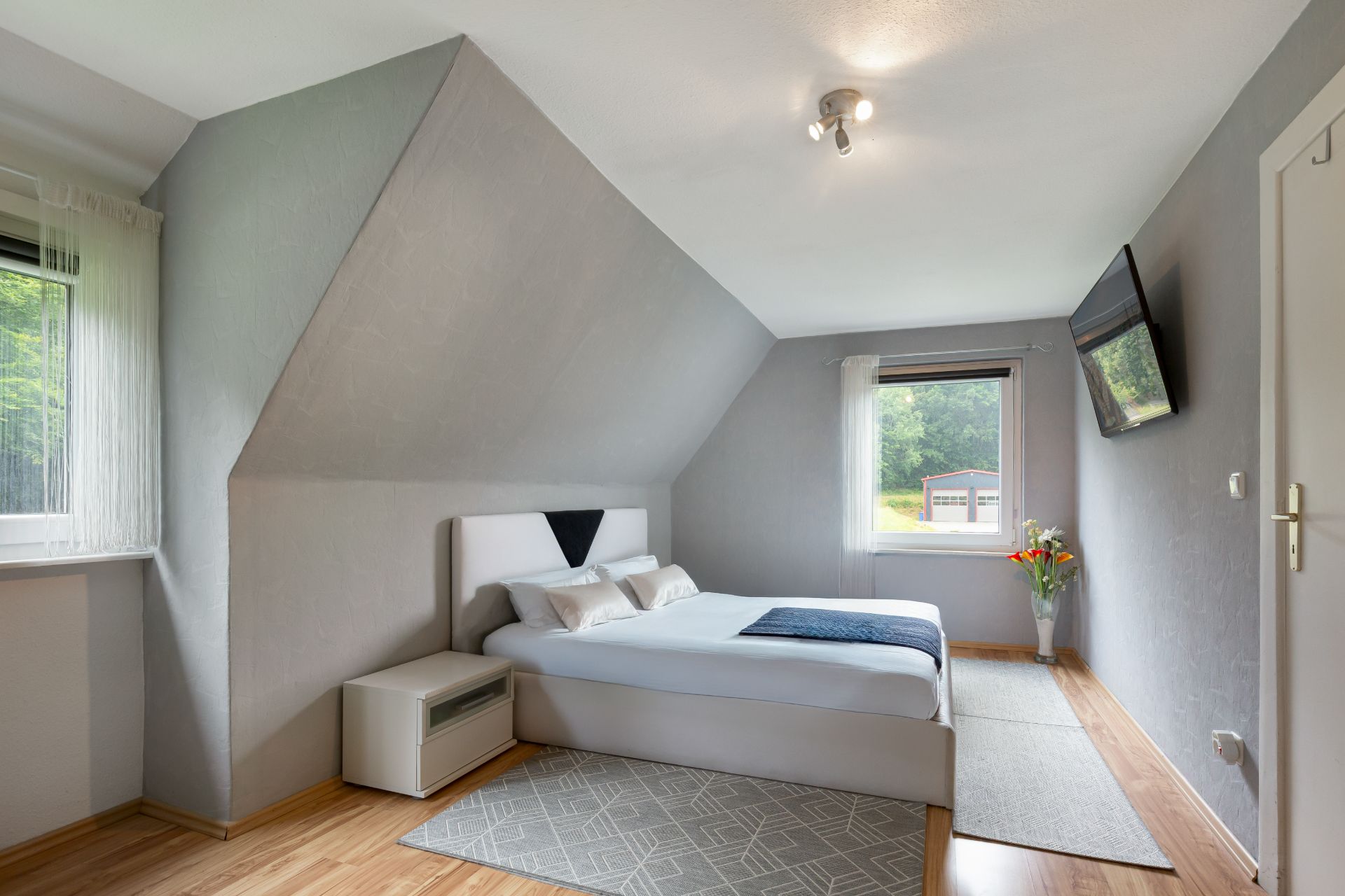 Modernes Schlafzimmer mit Doppelbett, Nachttisch, Teppich und Wandfernseher, zwei Fenstern und Holzboden.
