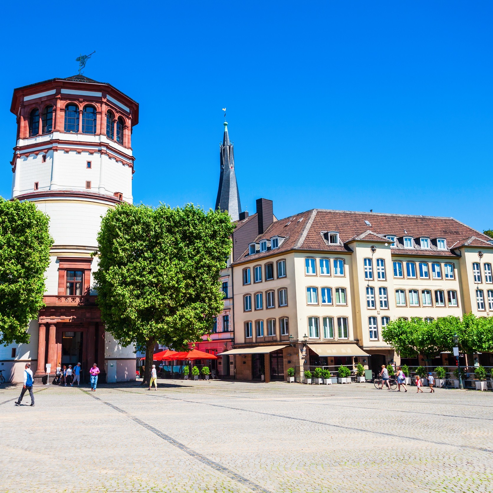 Wohnhaus mit mehreren Fenstern an einem sonnigen Platz, Bäume und einige Menschen sind sichtbar.