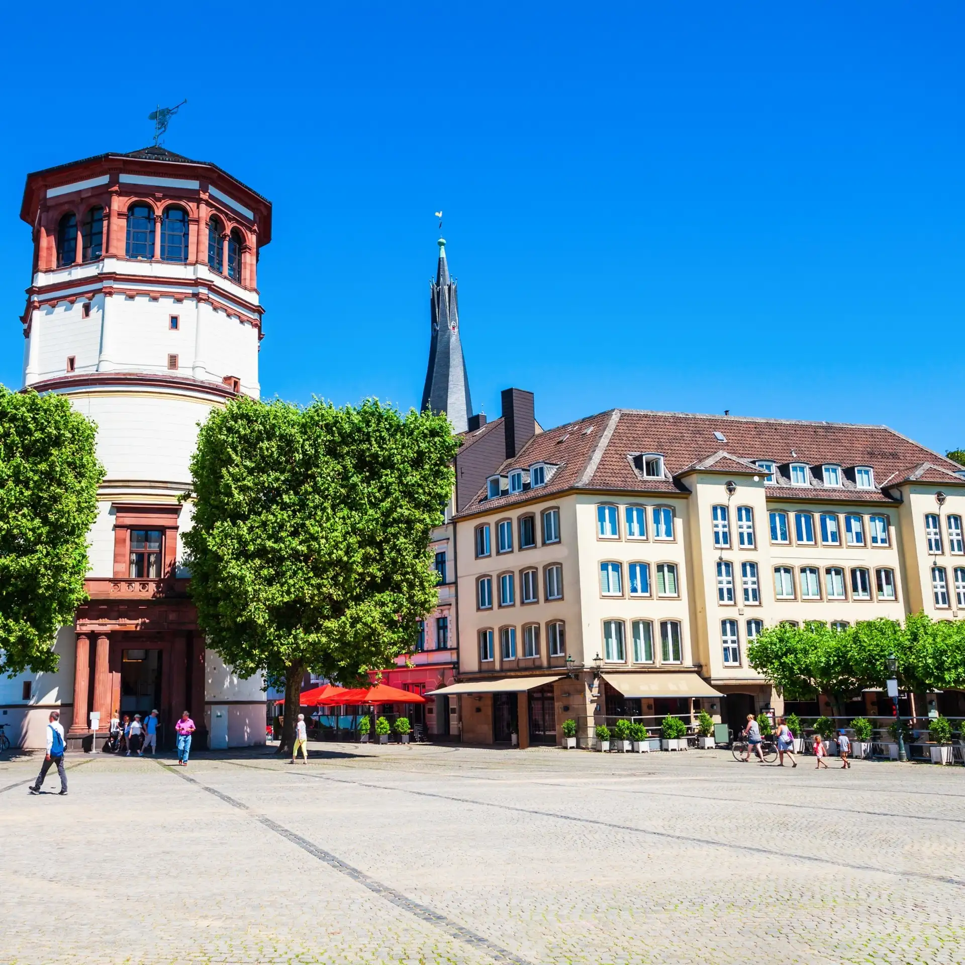 Wohnhaus mit mehreren Fenstern an einem sonnigen Platz, Bäume und einige Menschen sind sichtbar.