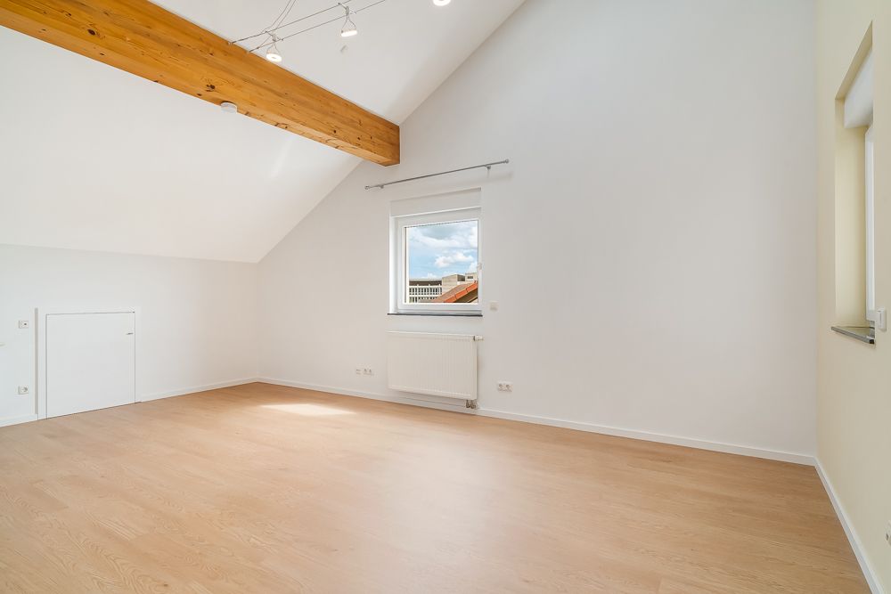Heller, leerer Raum mit Holzfußboden, Dachschräge, Fenster und sichtbarem Holzbalken an der Decke.