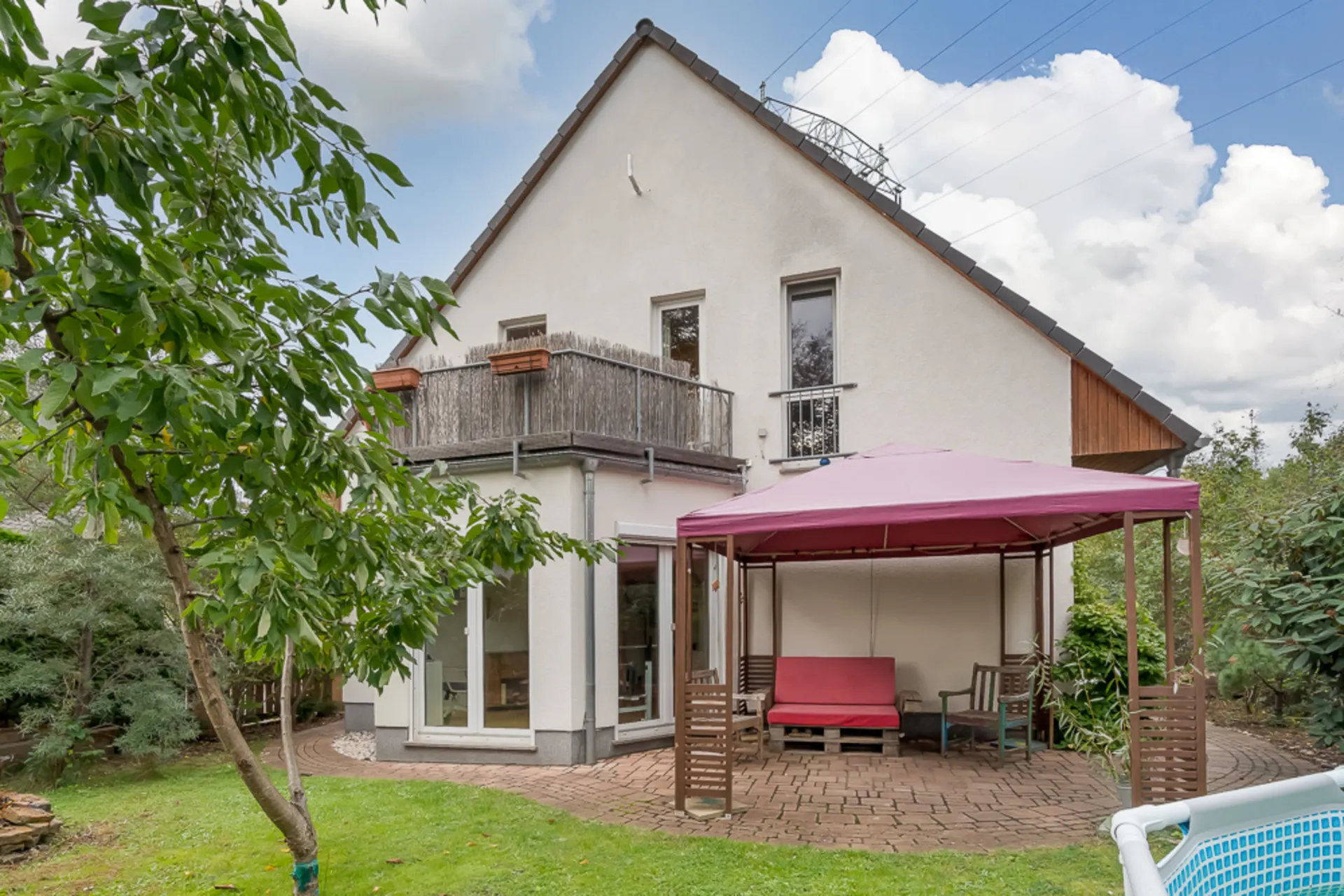 Zweistöckiges Haus mit Balkon, Garten, rotem Pavillon und Gartenmöbeln auf einer Terrasse.