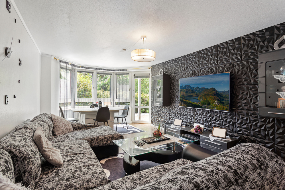 Modernes Wohnzimmer mit Ecksofa, Glastisch, TV an 3D-Wand und großem Fenster mit Esstisch.