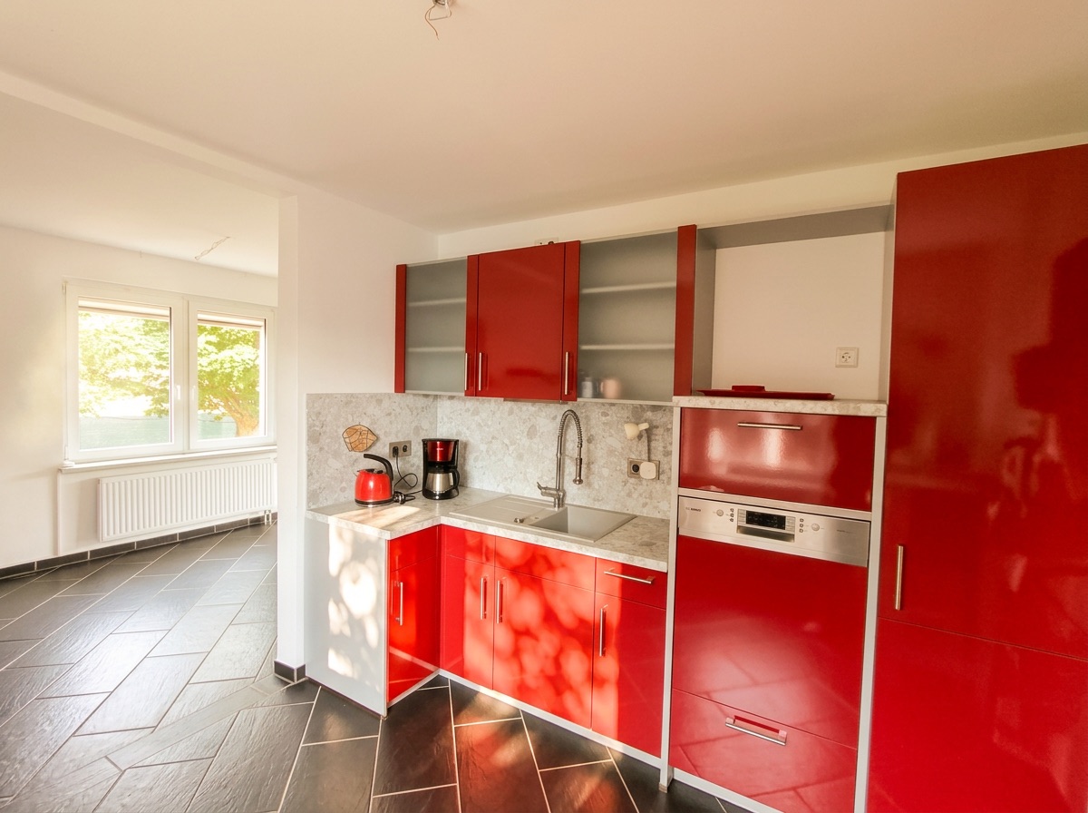 Moderne Küche mit roten Hochglanzschränken, Spüle und Geräten, angrenzend an einen hellen Raum mit Fenstern.