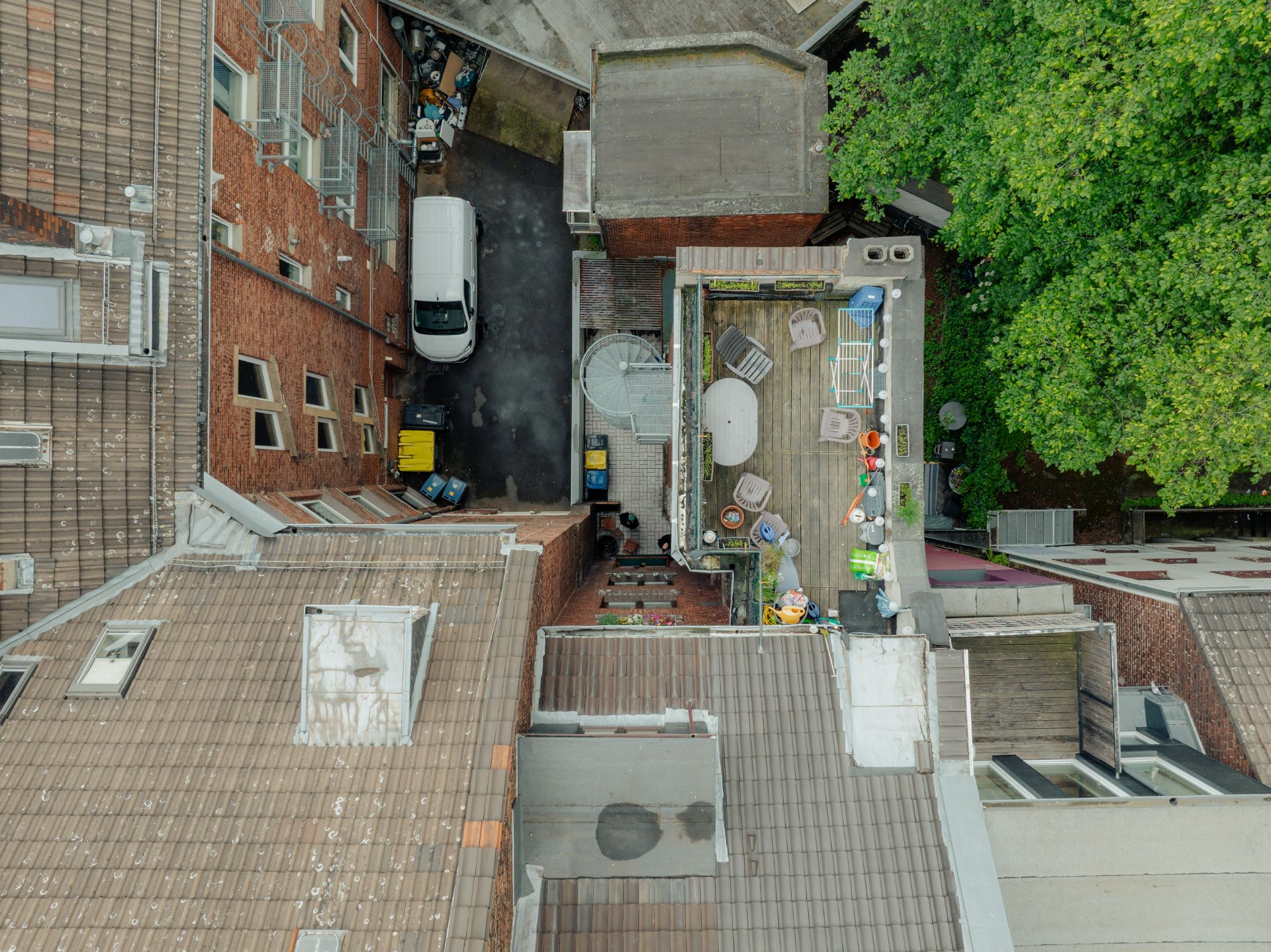 Dachterrasse mit Möbeln und Pflanzen, daneben Innenhof mit geparktem weißen Transporter, Bäume rechts.