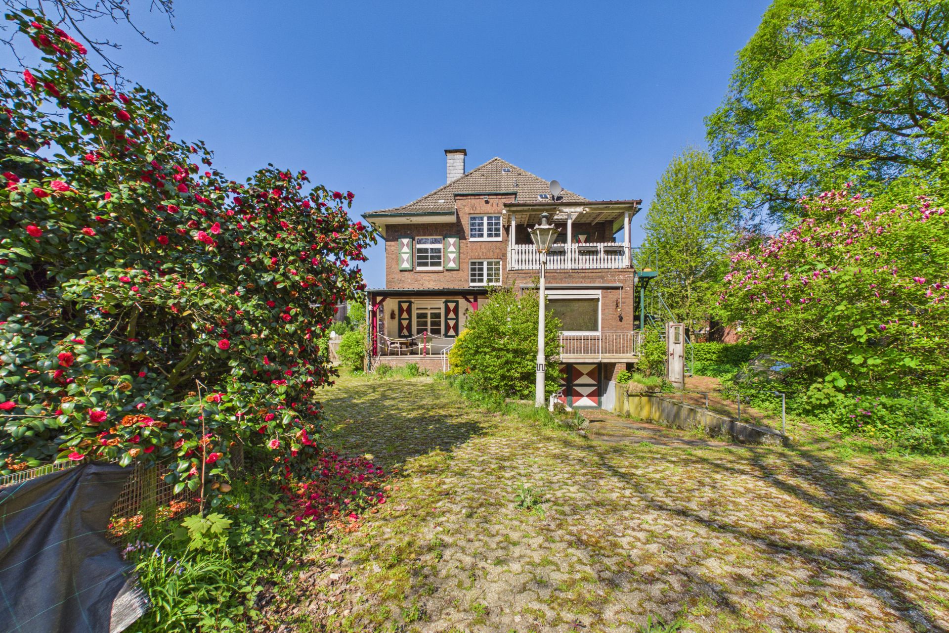 Großes Backsteinhaus mit Balkon, umgeben von blühenden Sträuchern und Bäumen im sonnigen Garten.