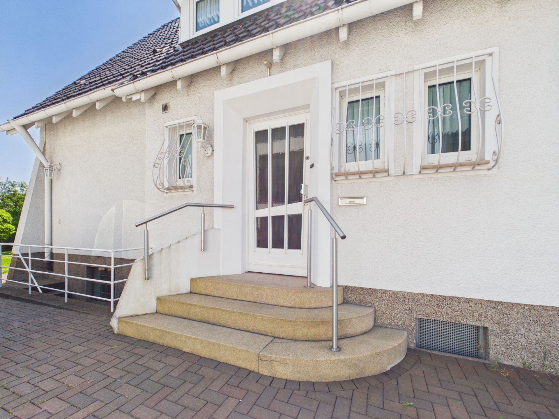 Weiße Haustür mit drei Stufen und Geländer, zwei vergitterte Fenster daneben.